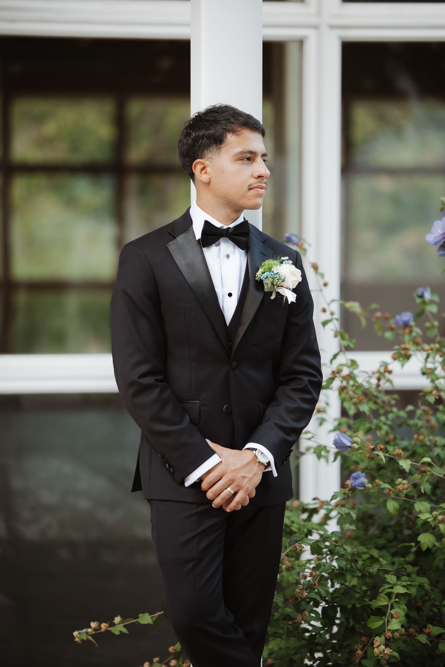 Groom in black tuxedo with boutonniere stands beside lush greenery outdoors.