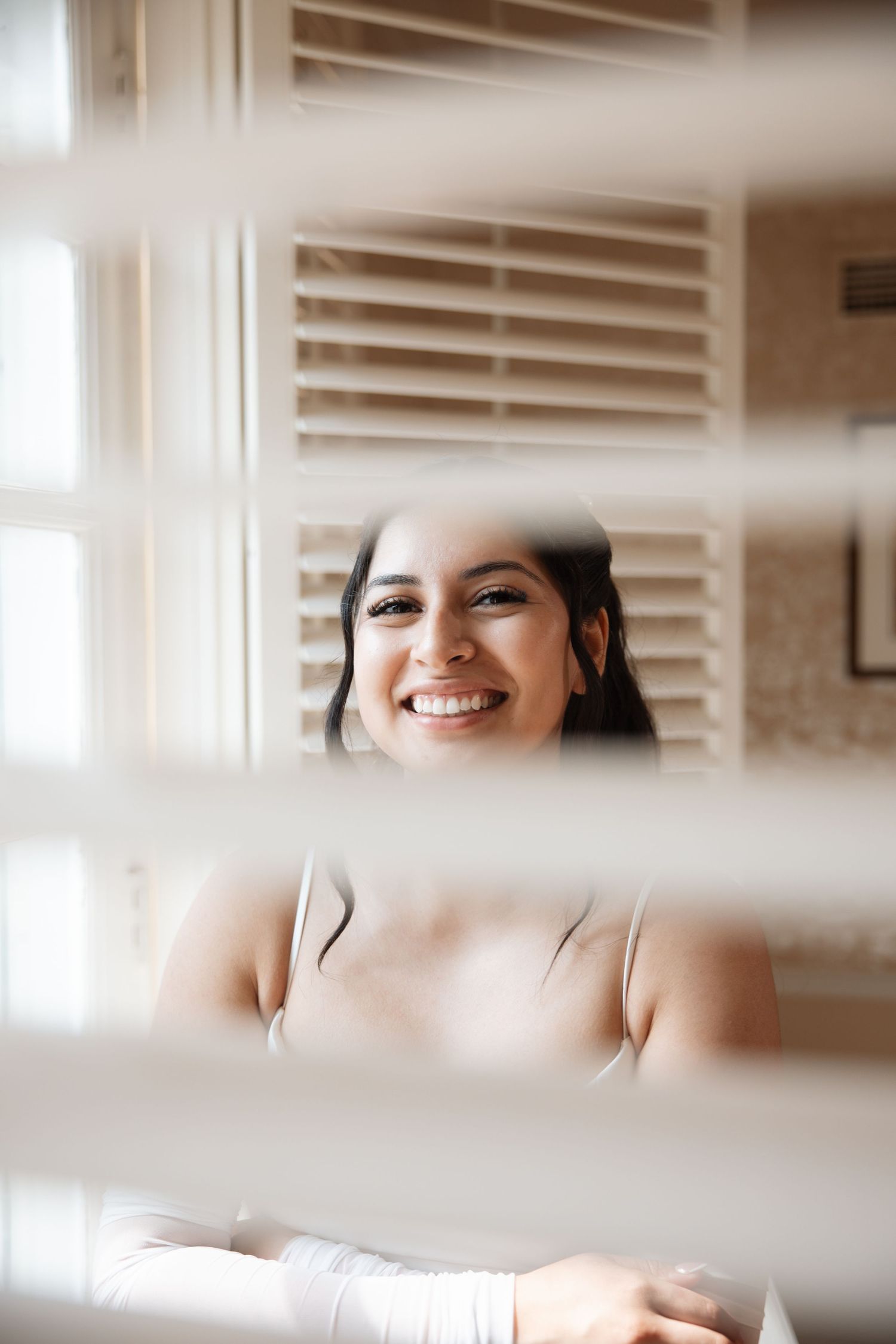 Joyful bride smiling beautifully as seen through white window blinds in soft natural light.