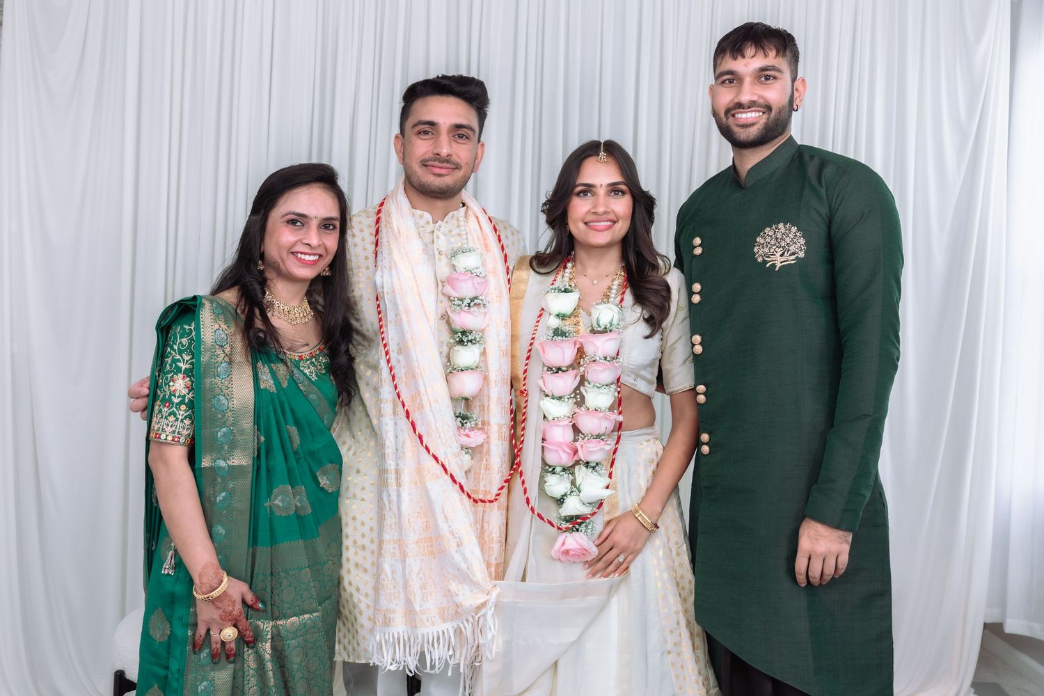 Couple in white wedding attire with floral garlands poses with family members in green and teal traditional Indian outfits.