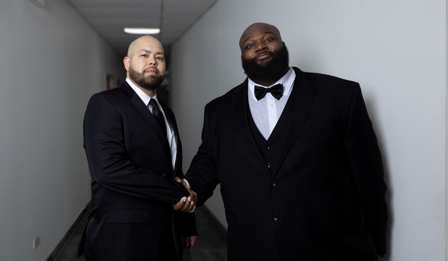 Two men in black tuxedos shaking hands in a bright hallway for a formal wedding portrait.