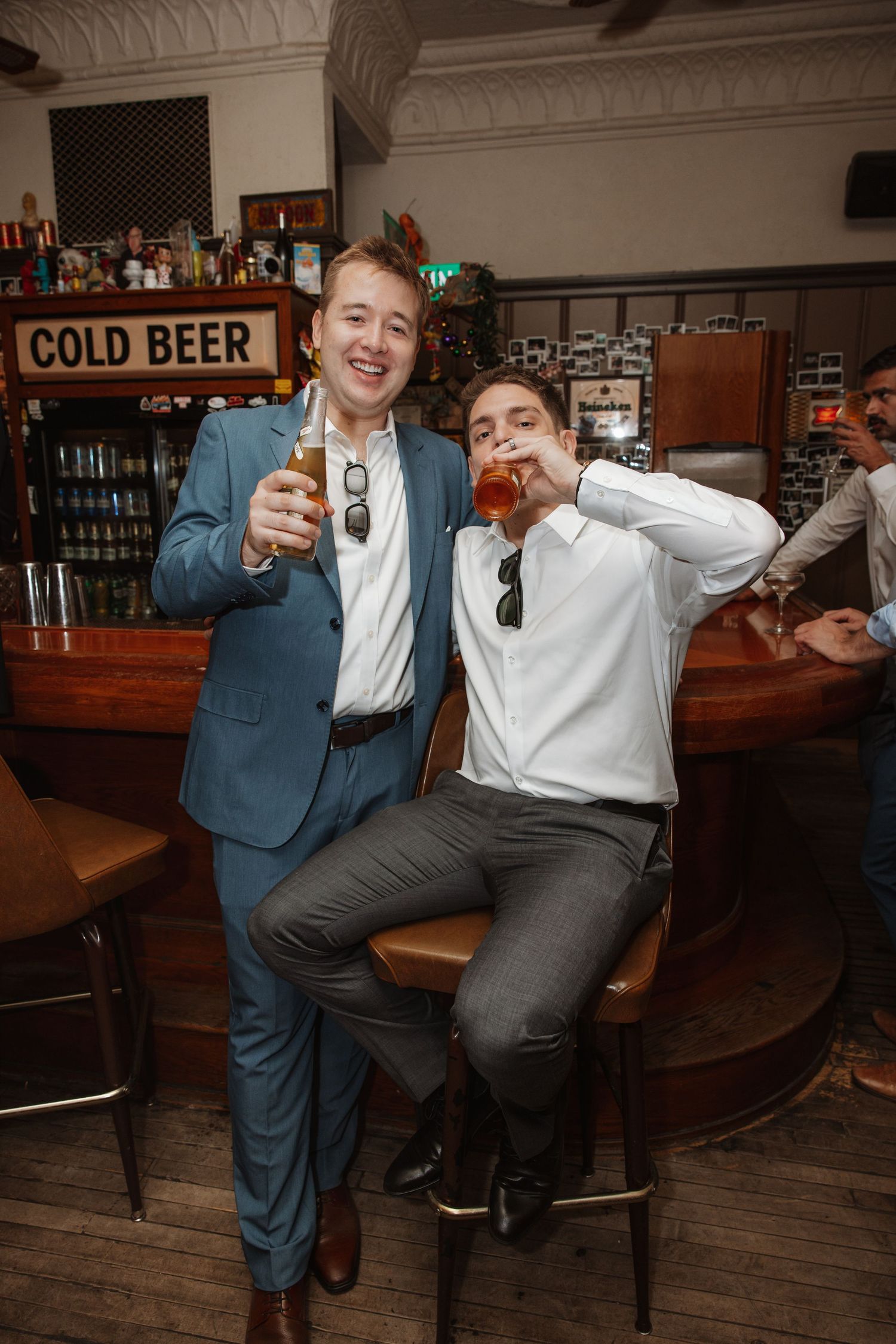 Two men in formal attire toast with drinks at a wooden bar with Cold Beer sign.