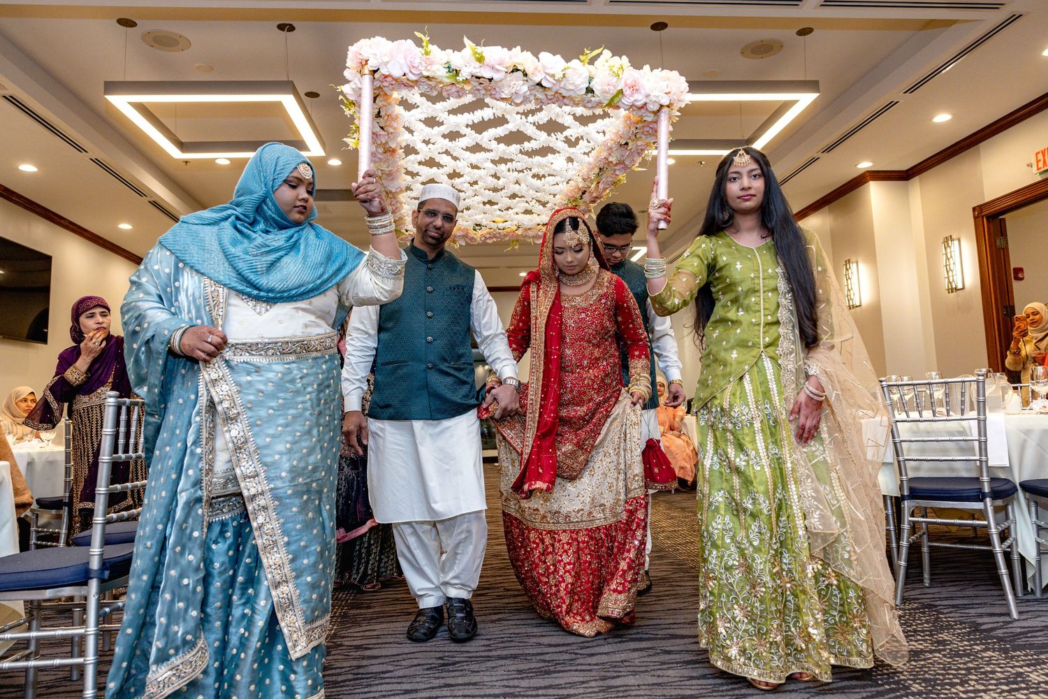 South Asian bride walks under flower canopy escorted by family in elegant traditional wedding attire.