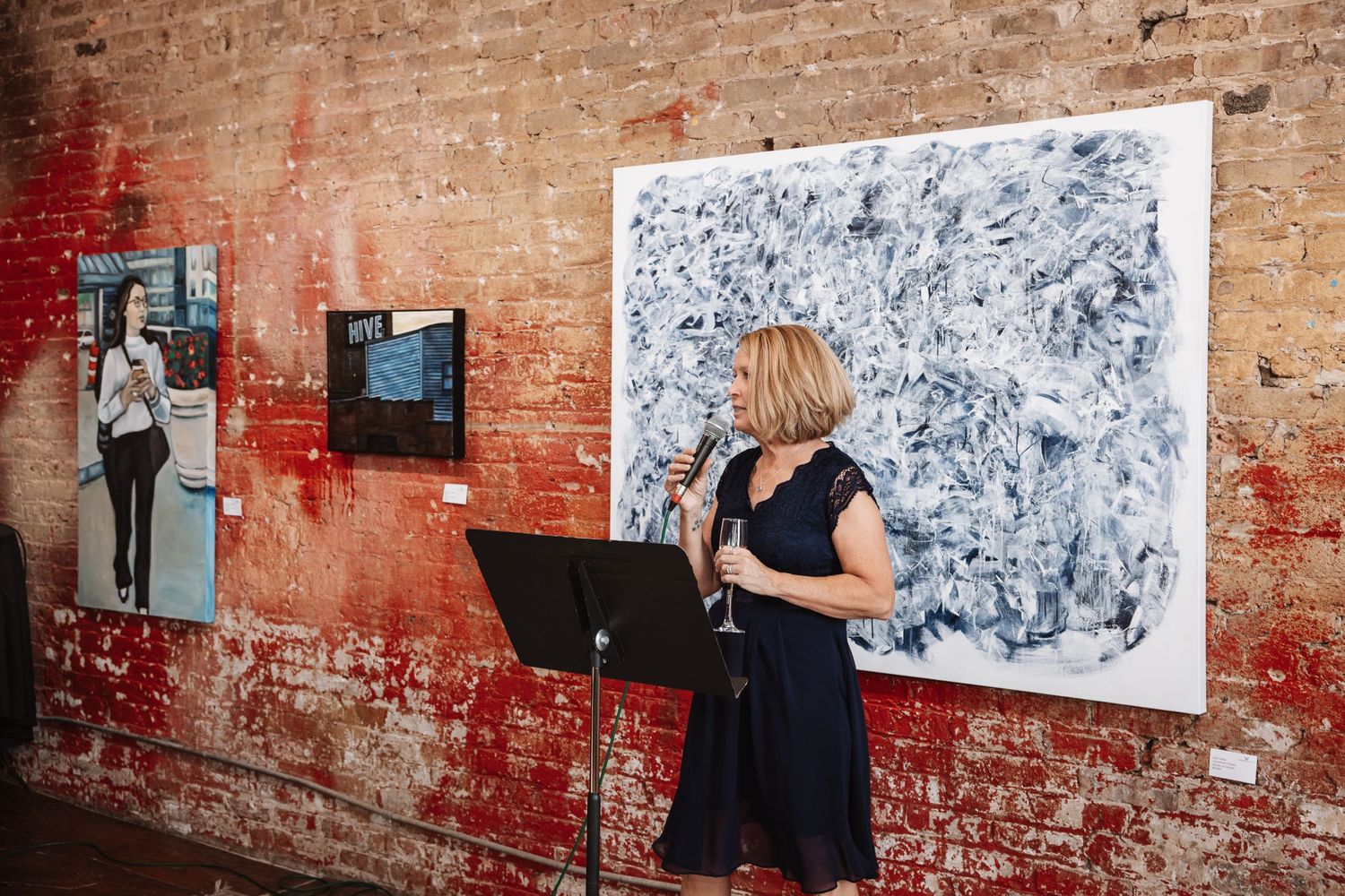 Woman in navy dress speaking at podium beside large abstract painting at an art gallery event.