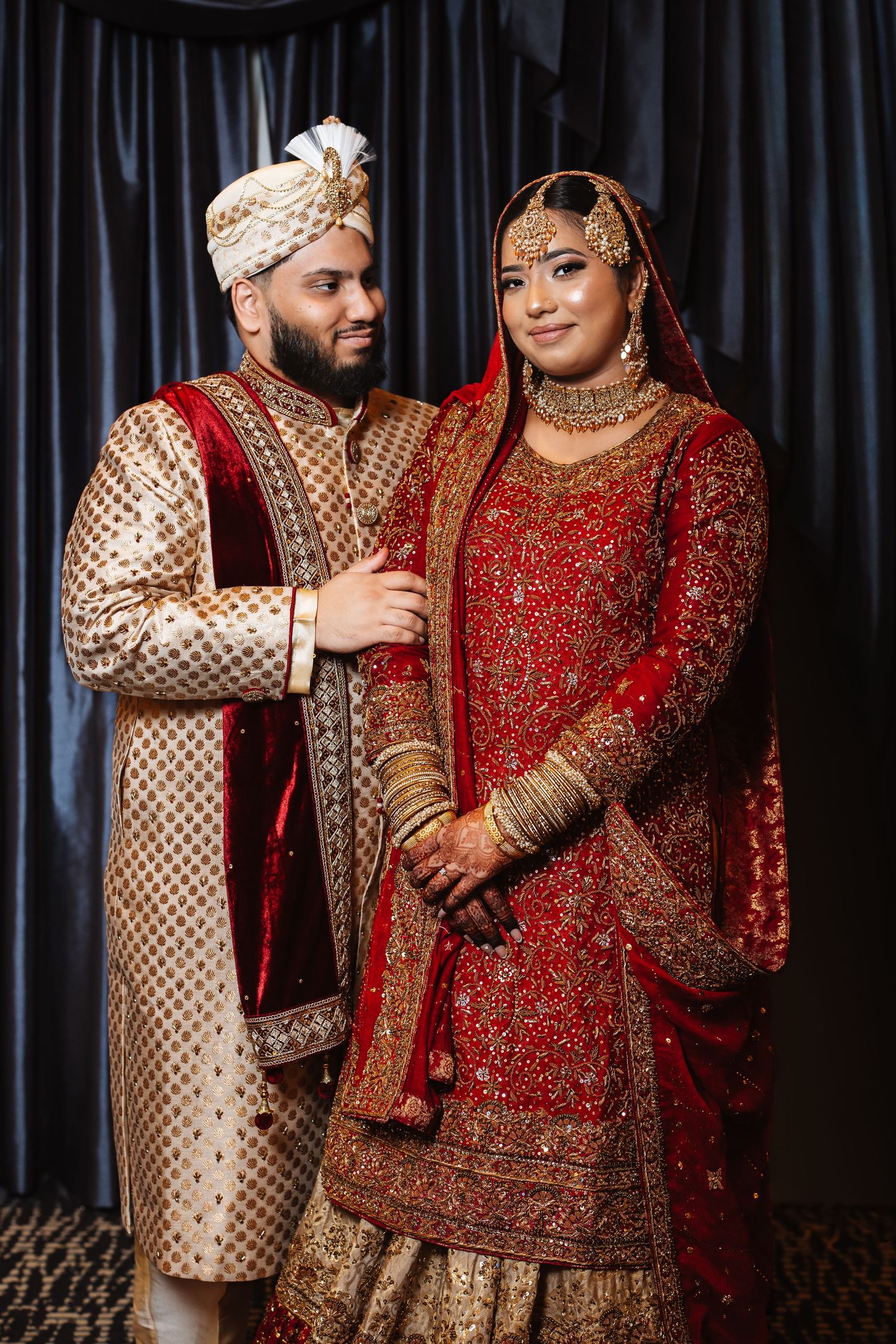 South Asian wedding couple in traditional embroidered bridal wear posing for formal portrait indoors.