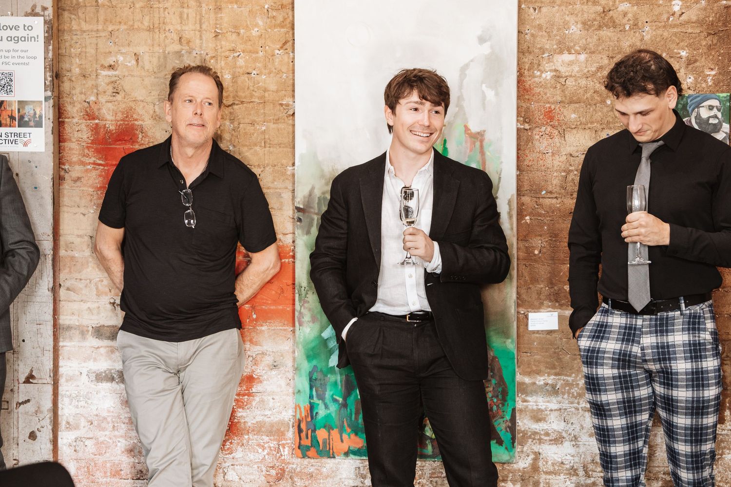 Three men in smart casual attire stand together at an art gallery social event.