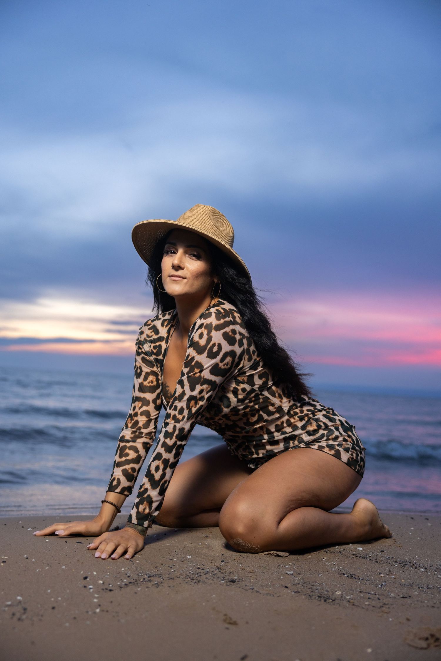 Woman in leopard print dress and wide brim hat kneeling on beach at dusk.