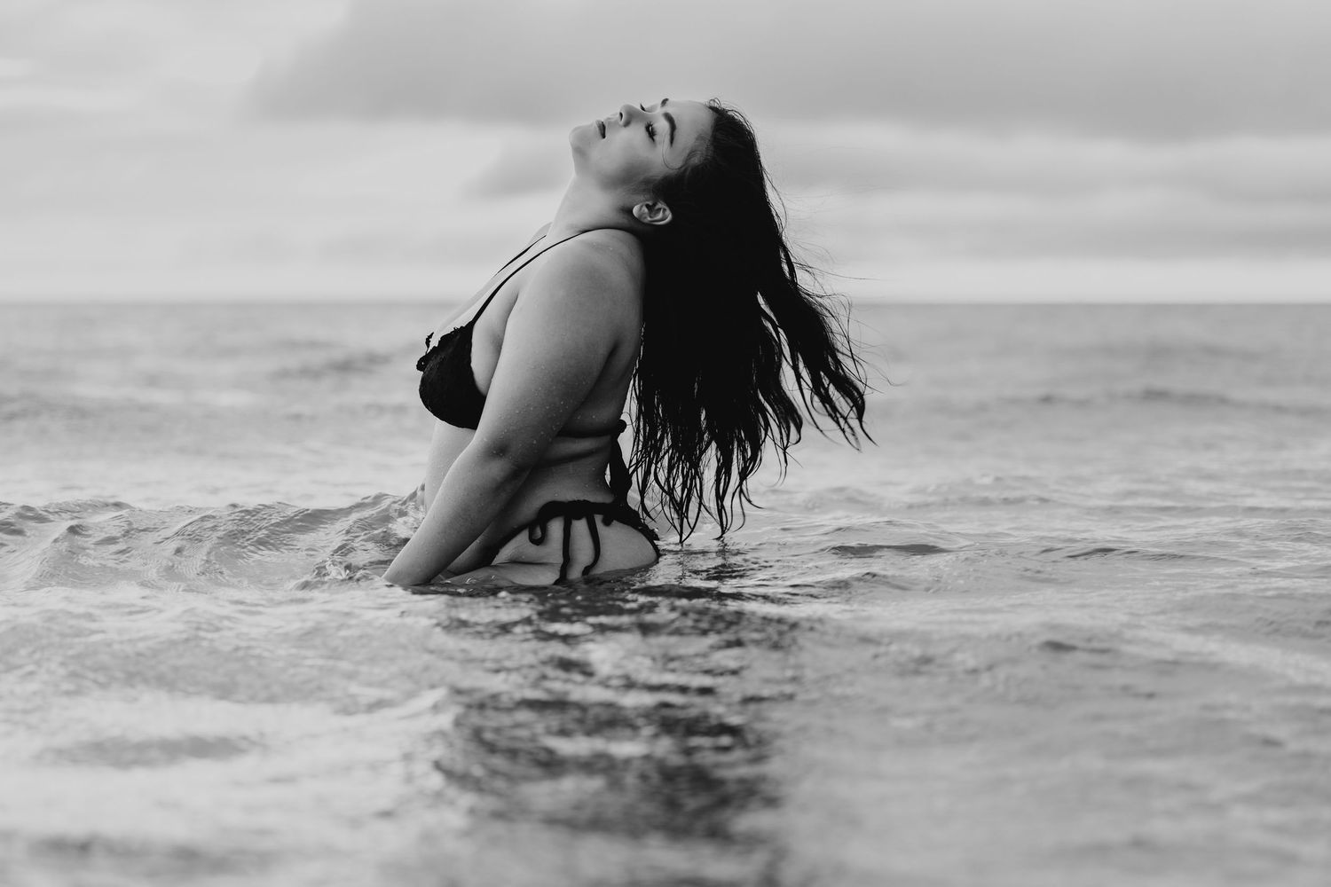 Black and white photo of woman in bikini tossing wet hair in ocean waves.