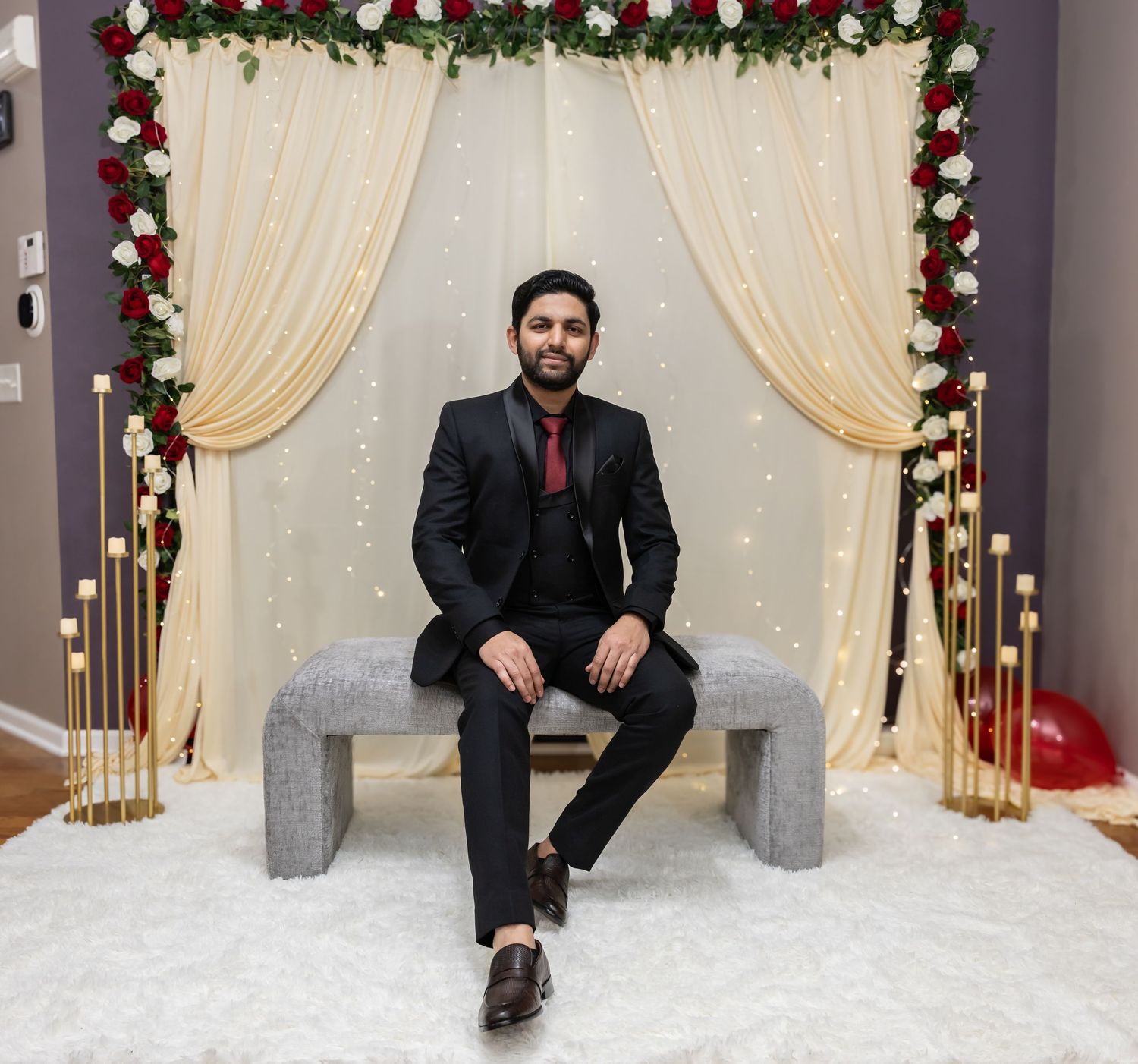 A groom in a black suit sits on a bench at a beautifully decorated wedding altar with red and white flowers.