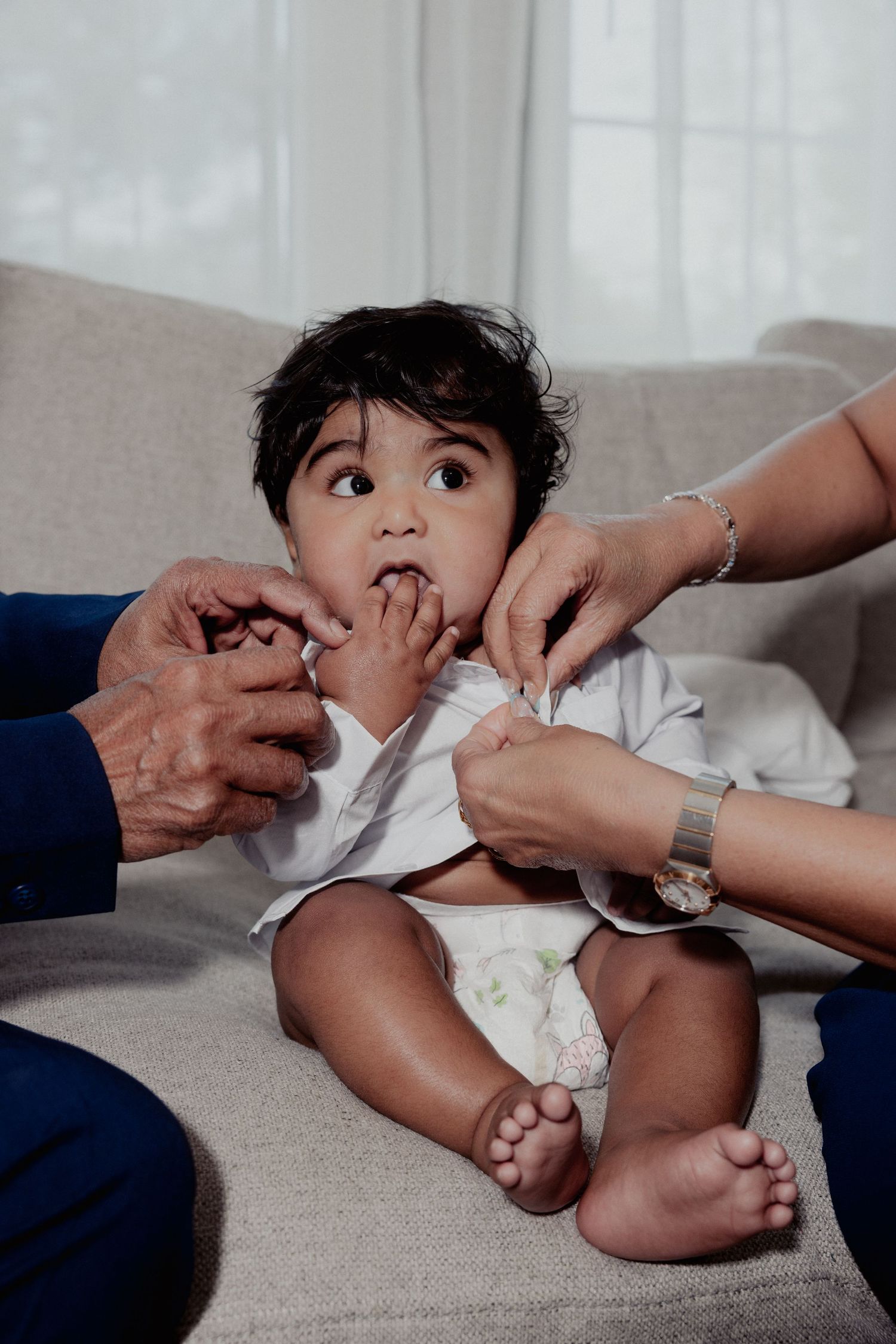 Baby in white shirt sitting on sofa being dressed by family hands for baptism.
