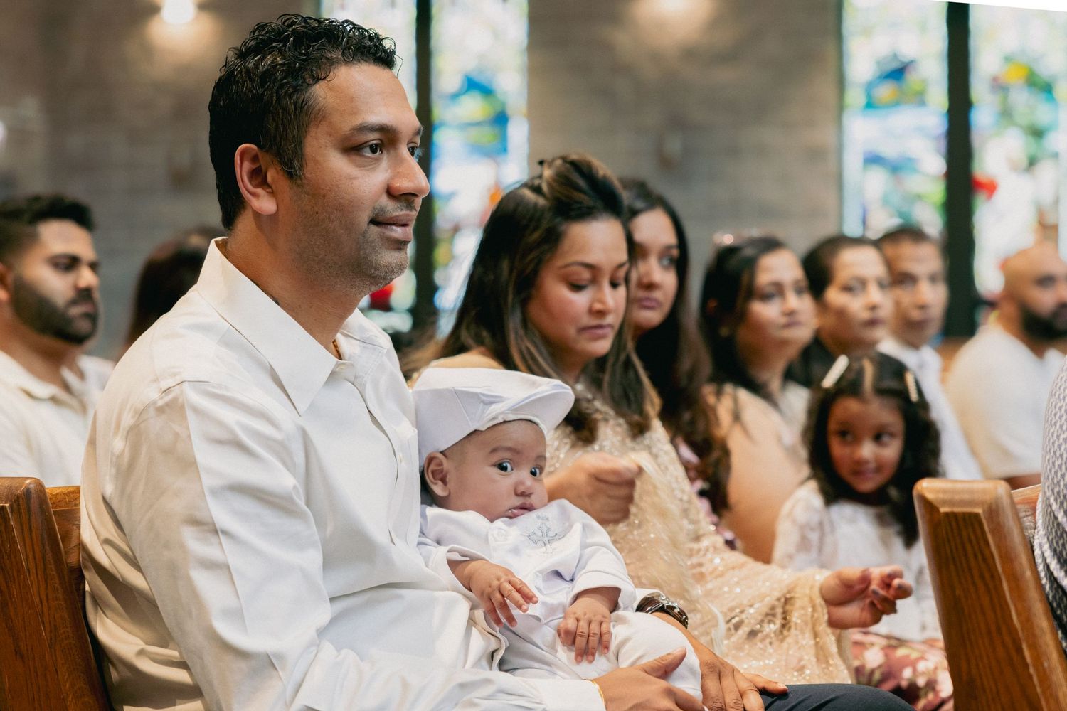 Asian man holds baby in white baptism outfit while seated in church pew.