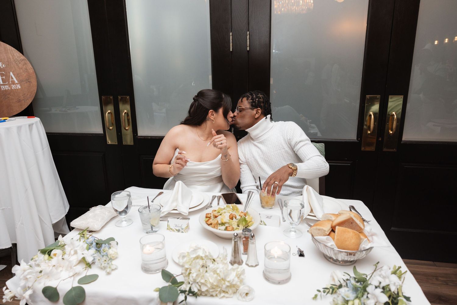 Bride and groom share romantic kiss at sweetheart table decorated with white flowers during reception.