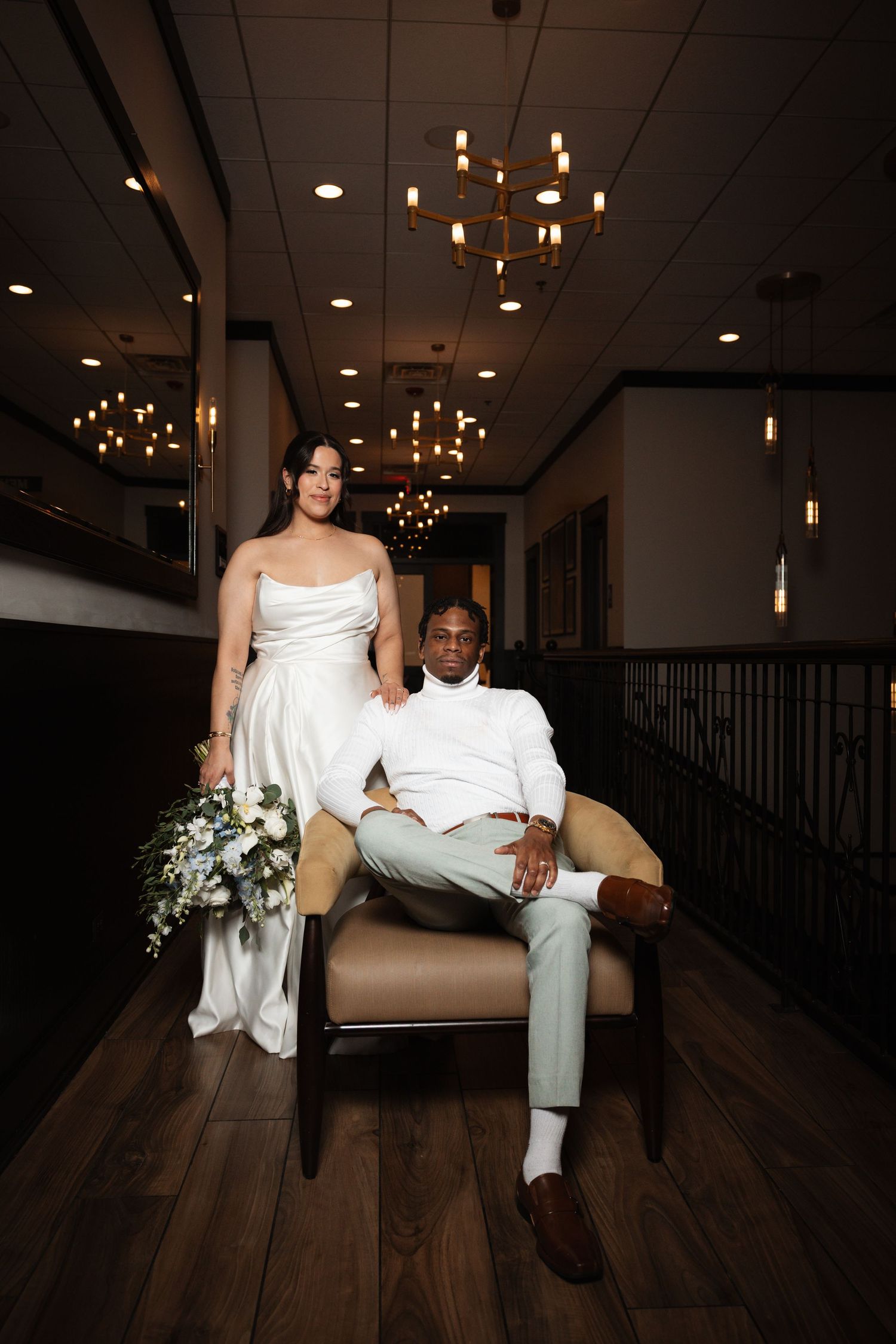 Wedding couple posing in elegant venue, bride holding bouquet beside seated groom.