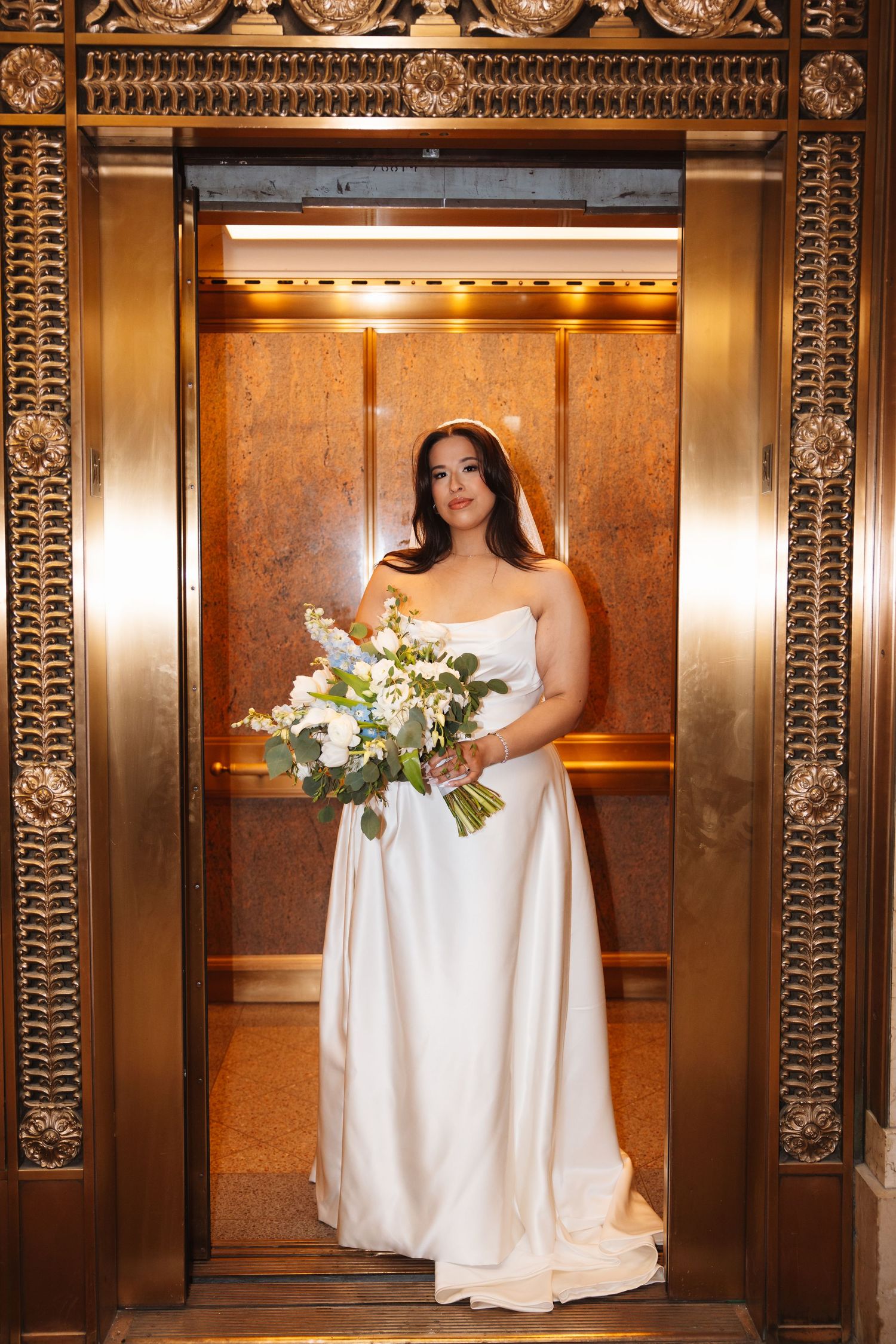 Bride in strapless white gown holding floral bouquet stands in ornate gold elevator.