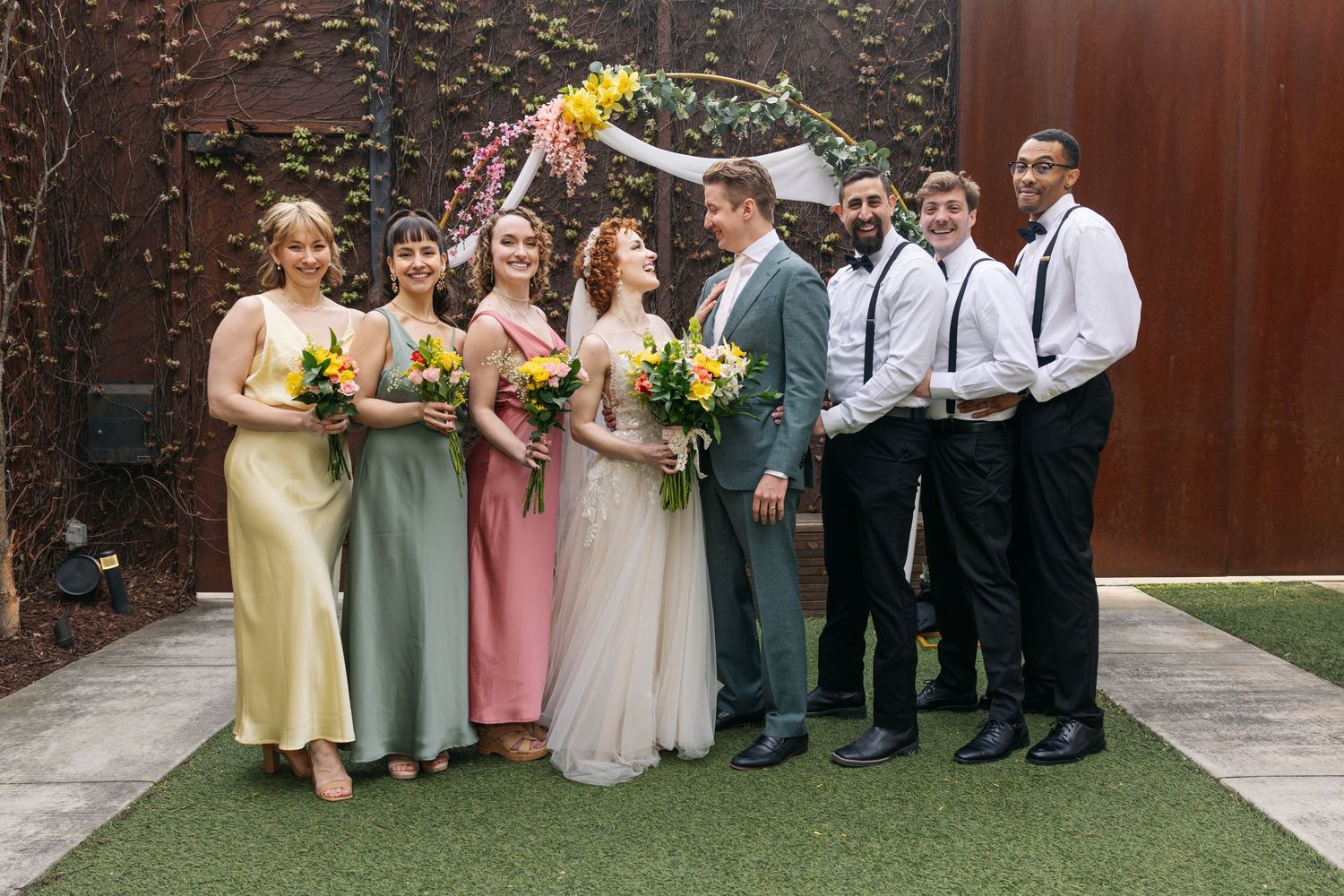 Wedding party poses together outdoors, bridesmaids in colorful gowns and groomsmen in suits with suspenders.