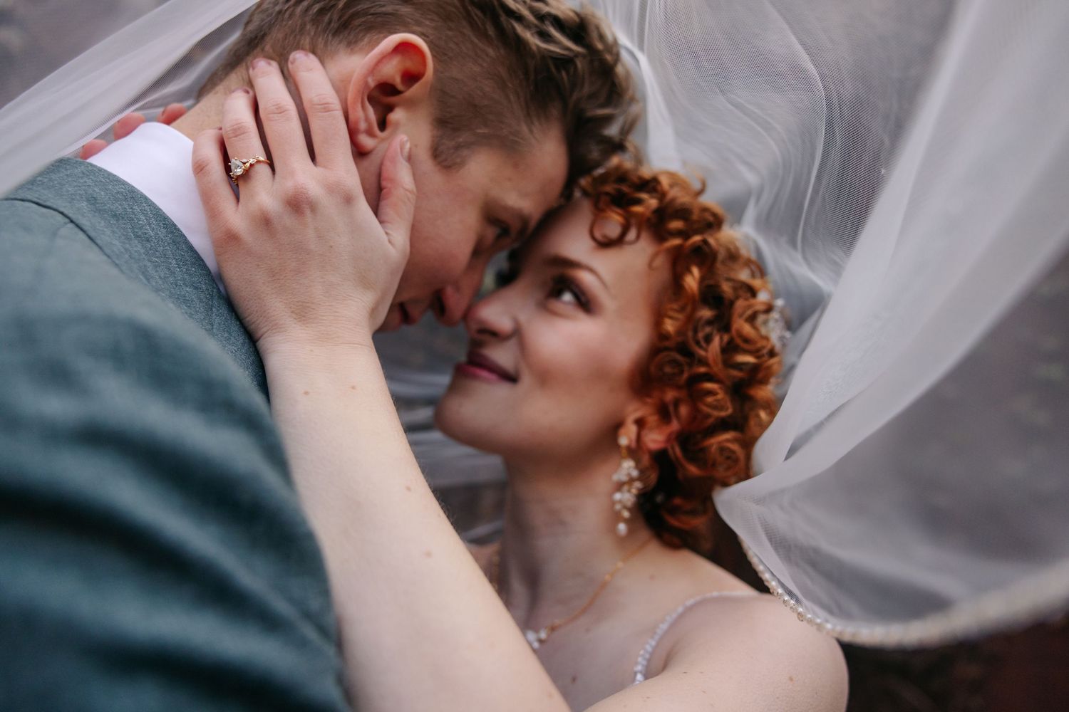 Bride and groom share an intimate moment under a wedding veil, with the bride smiling softly at the camera.