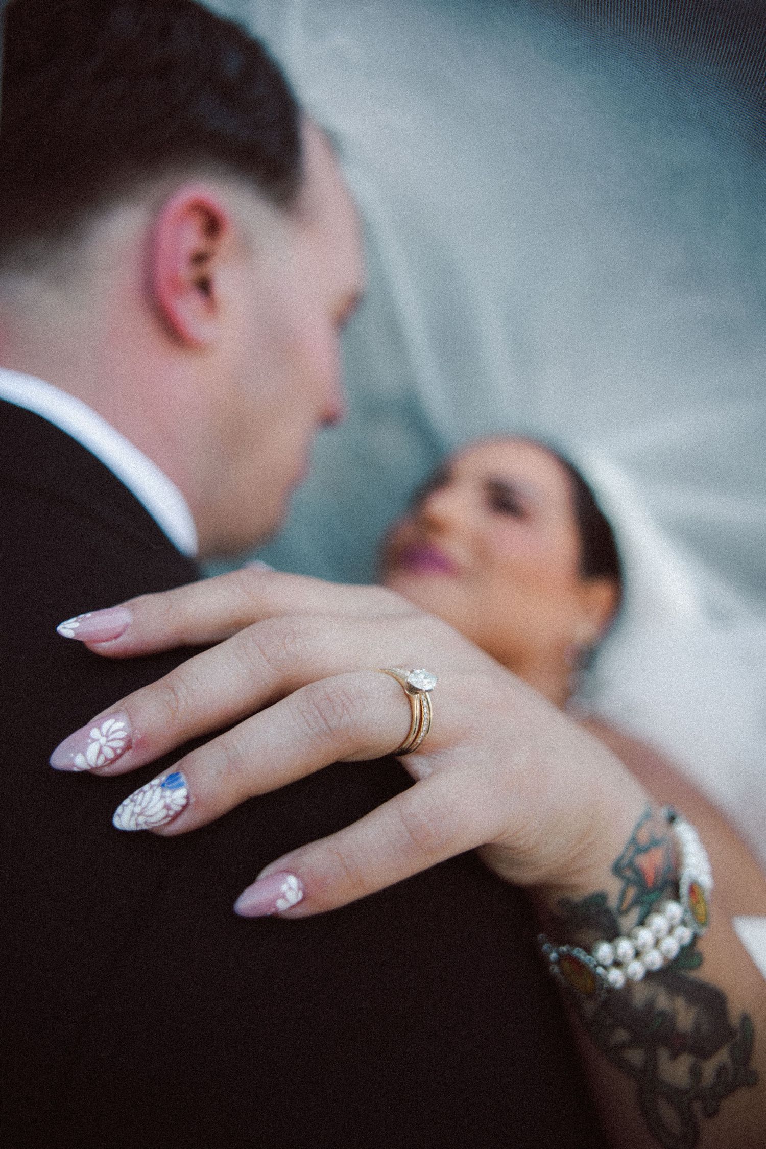 Close-up of bride's hand with gold diamond engagement ring and floral nail art resting on groom's shoulder.