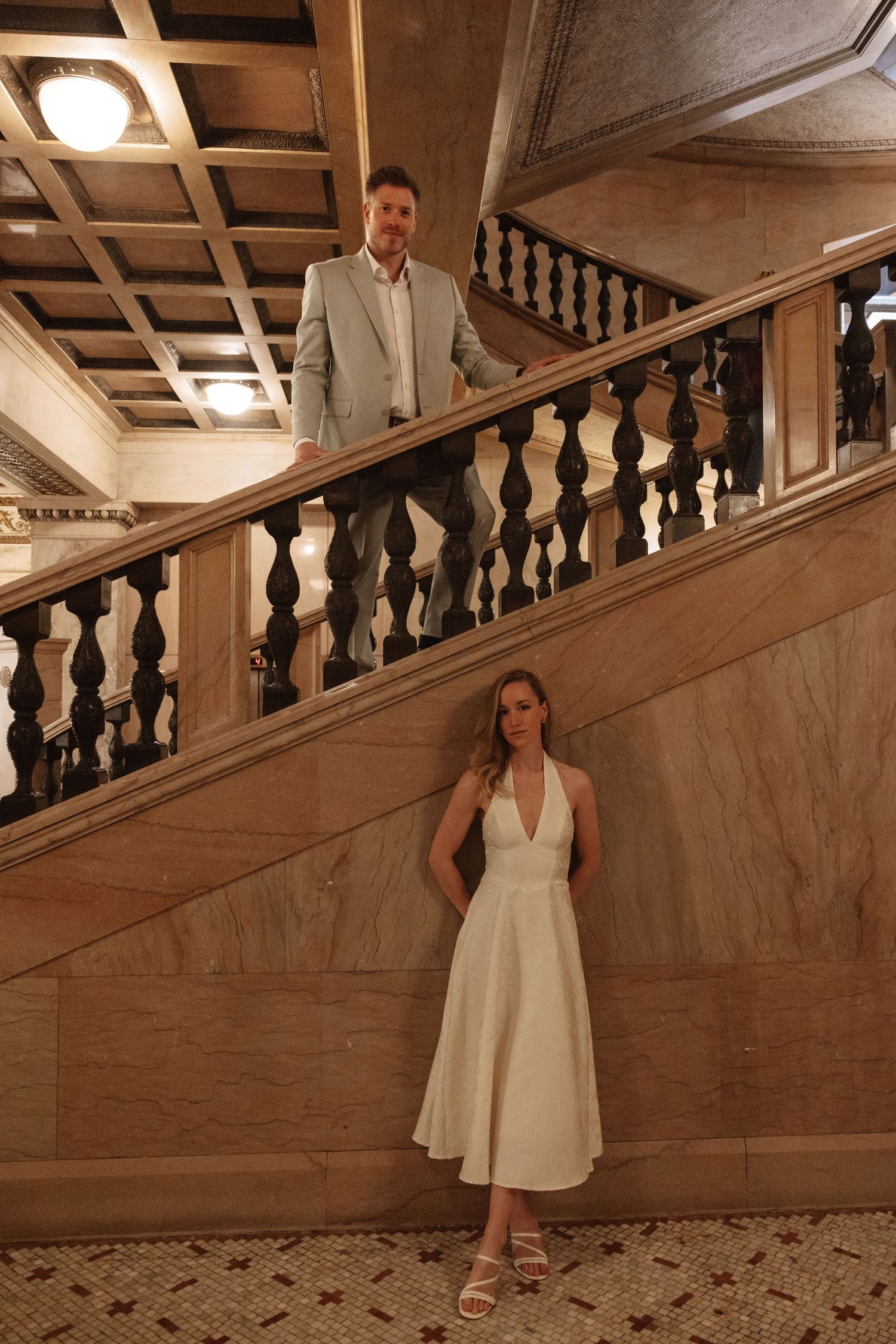 Groom in light suit stands on grand staircase above bride in white halter dress in elegant hall.