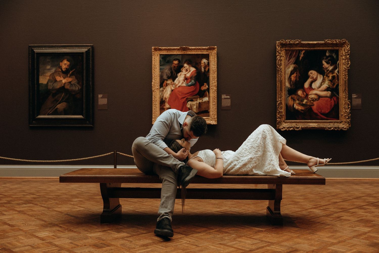 Couple sharing a romantic kiss on a museum bench surrounded by classic paintings in an art gallery.