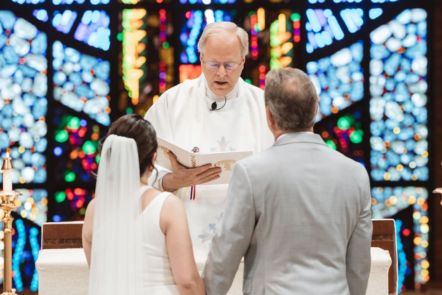 Officiant reading vows to bride and groom at colorful stained glass church wedding ceremony altar.