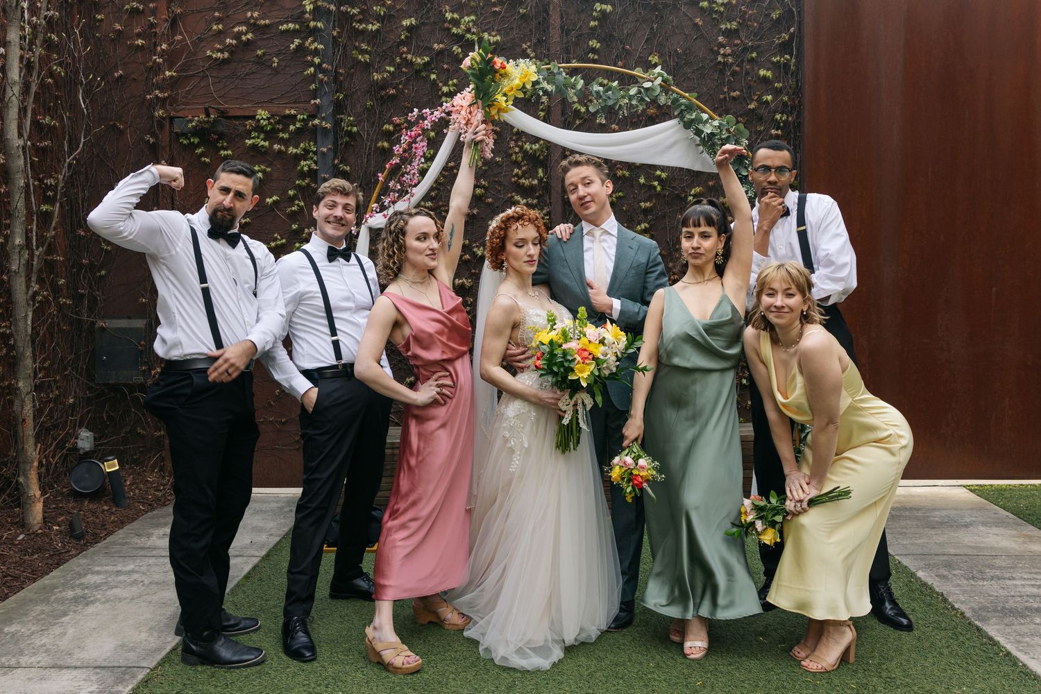 Wedding party group photo with bride, groom, bridesmaids in colorful dresses and groomsmen celebrating outdoors.