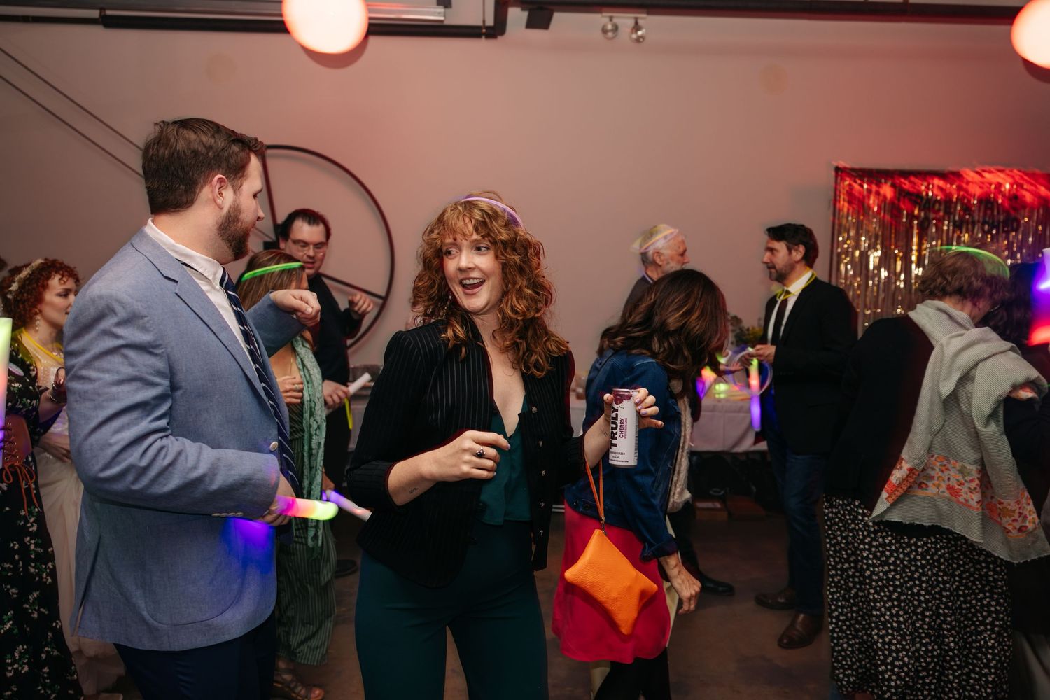 Red-haired woman in green velvet outfit laughs and socializes with guests at a lively wedding reception party.