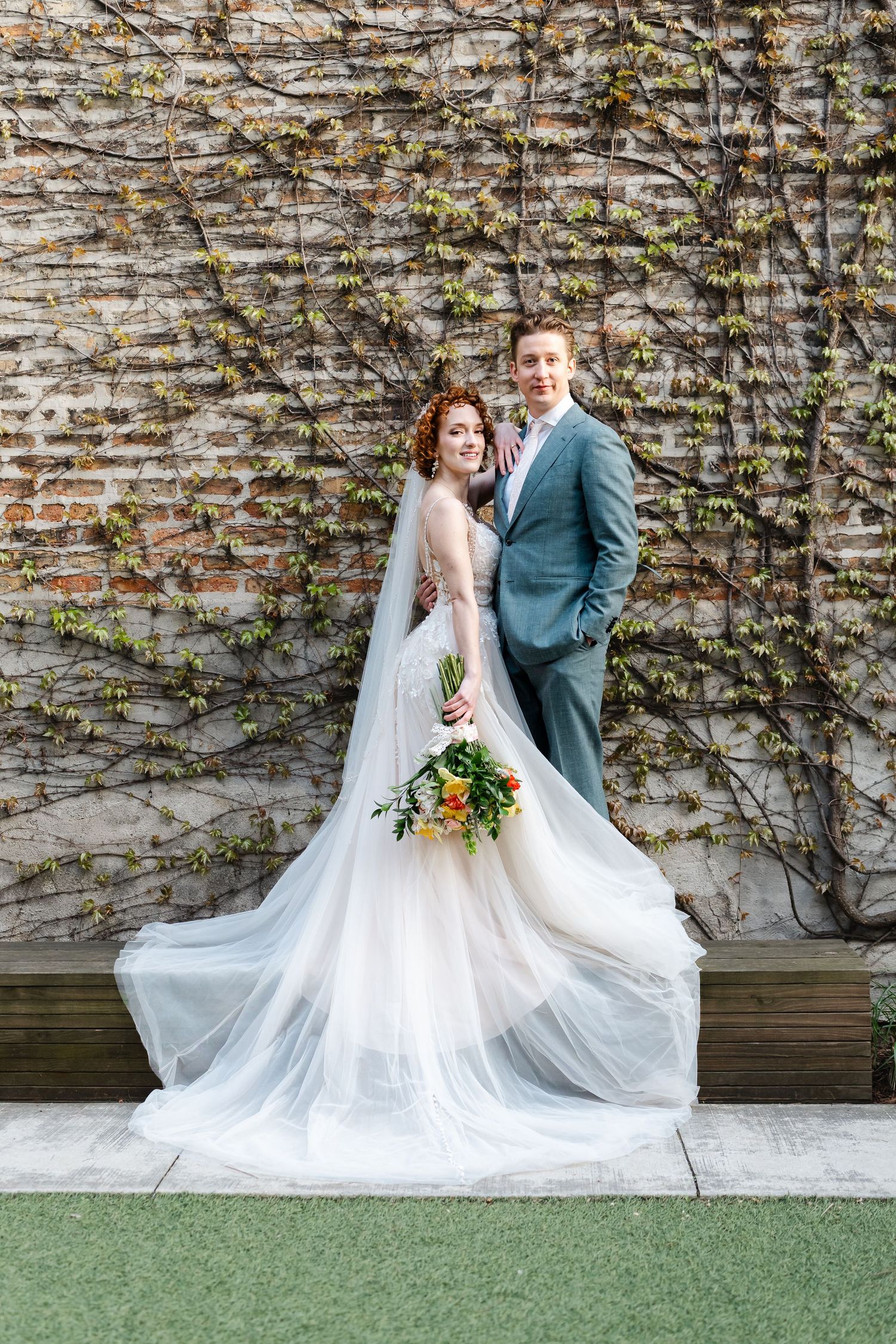 Bride in flowing white tulle gown with colorful bouquet and groom in teal suit pose before ivy-covered brick wall.