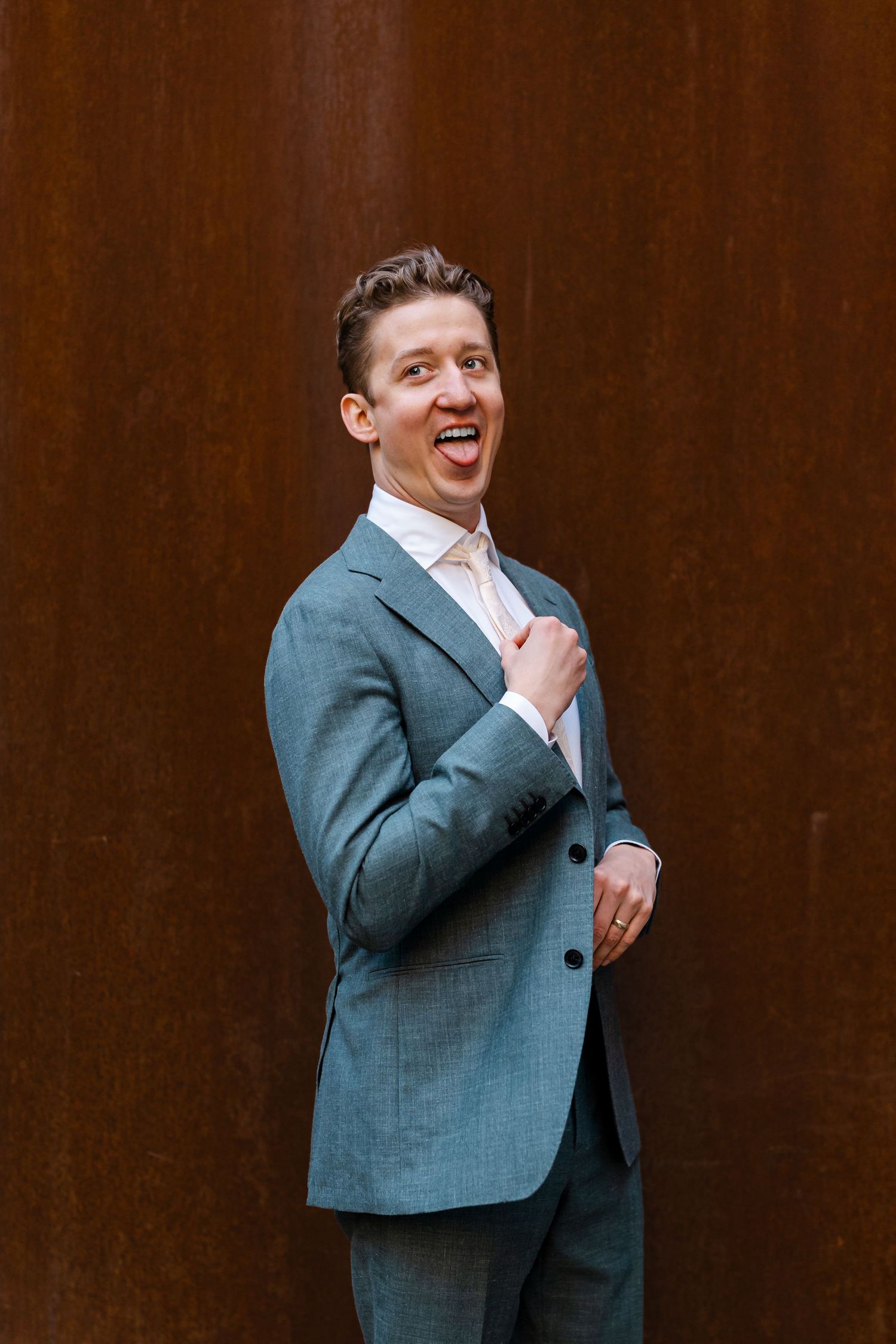 Groom in teal suit laughing joyfully while standing against rustic brown metal wall backdrop.