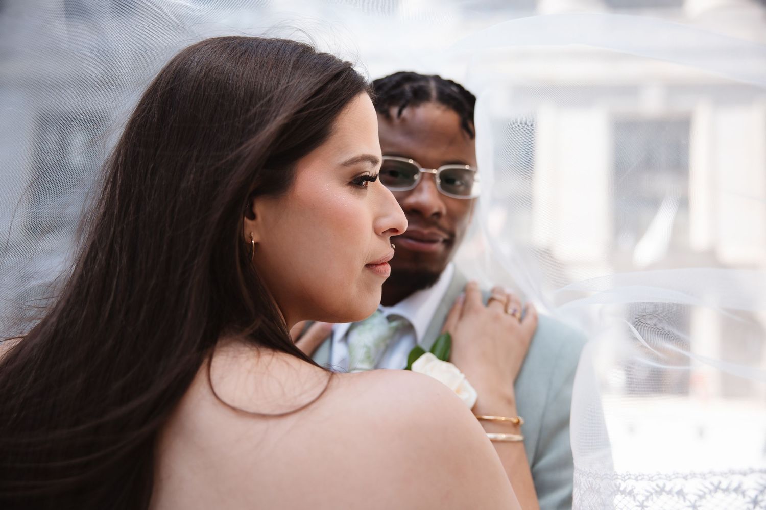 Bride and groom in elegant wedding attire, groom wearing sunglasses and boutonniere, bride with flowing veil.