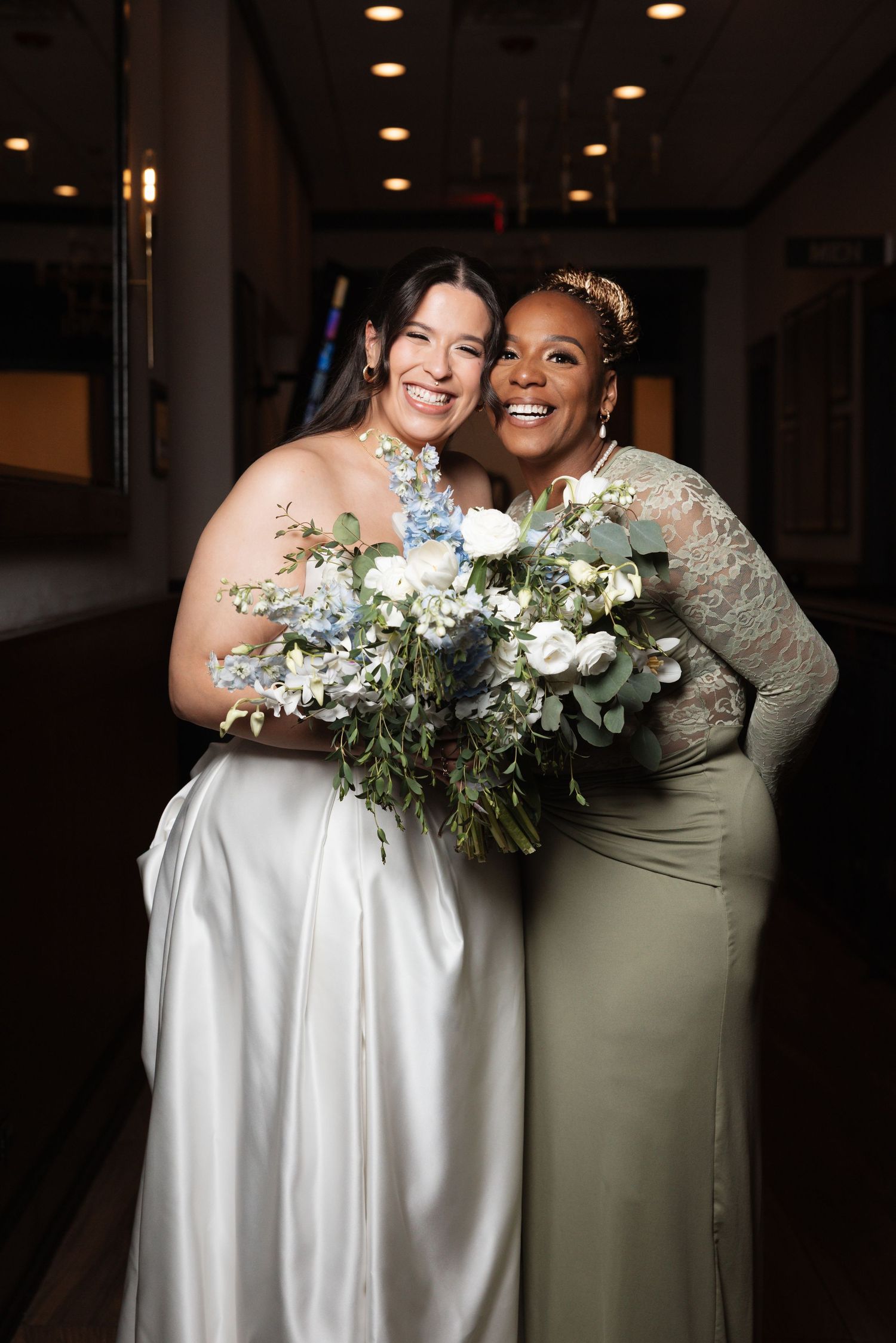 Bride and bridesmaid in sage green dress embrace holding blue and white bouquets.