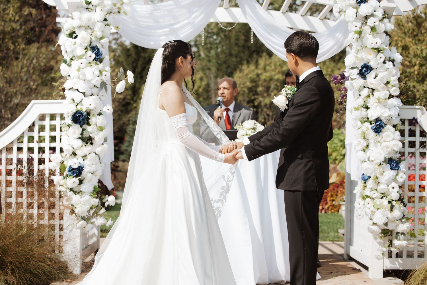 Bride in white gown and groom in black tuxedo hold hands at floral wedding arch ceremony.