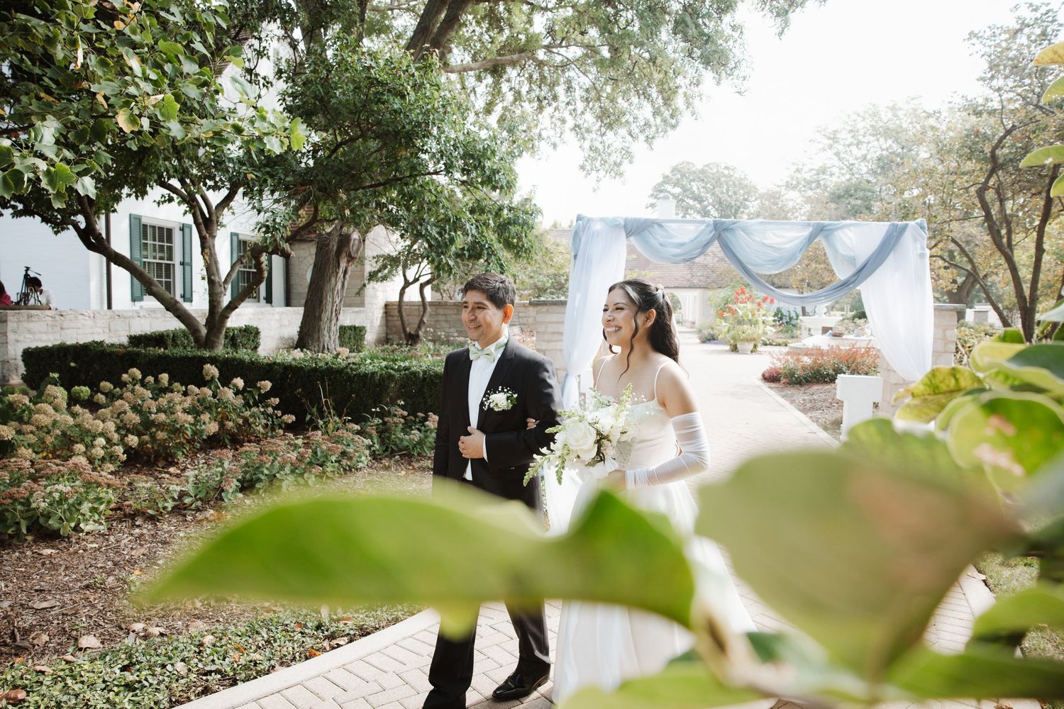 Bride in white gown and veil walks with groom through garden wedding venue.