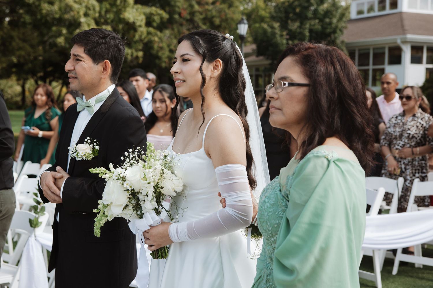 Bride walking down the aisle escorted by parents during an outdoor garden wedding ceremony.