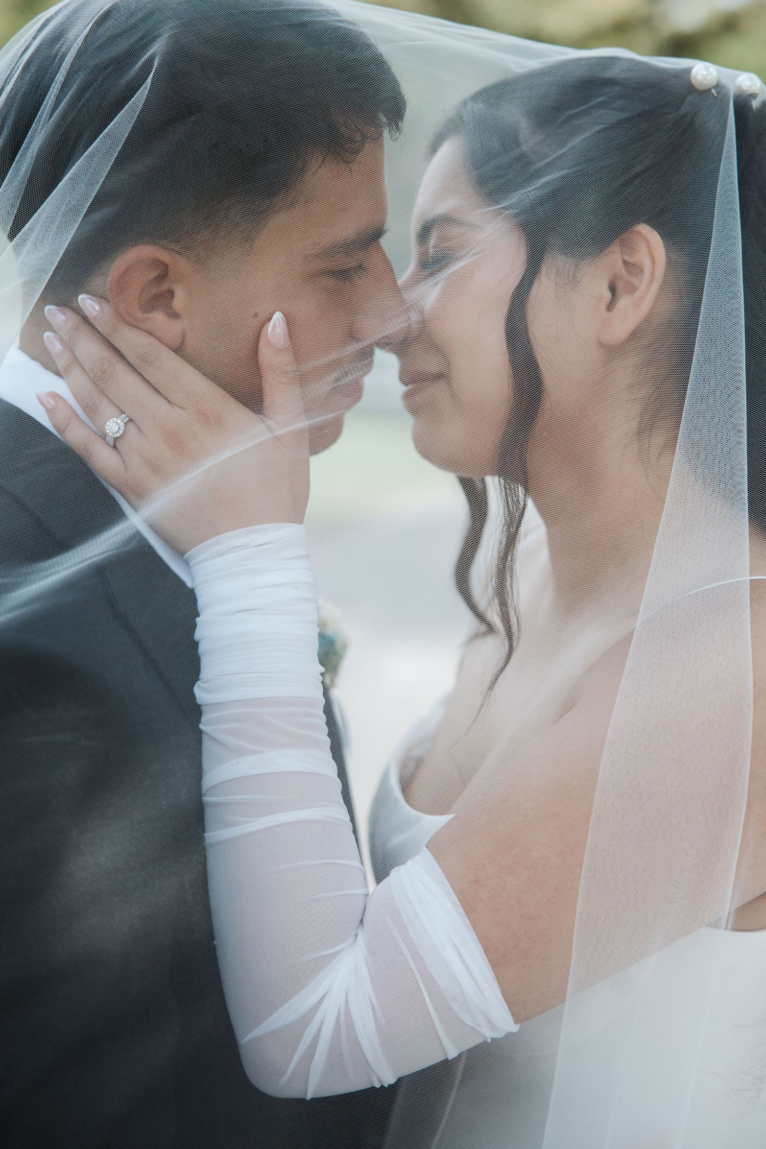 Newlyweds tenderly touch foreheads beneath a bridal veil, bride wearing engagement ring and elegant white gown.
