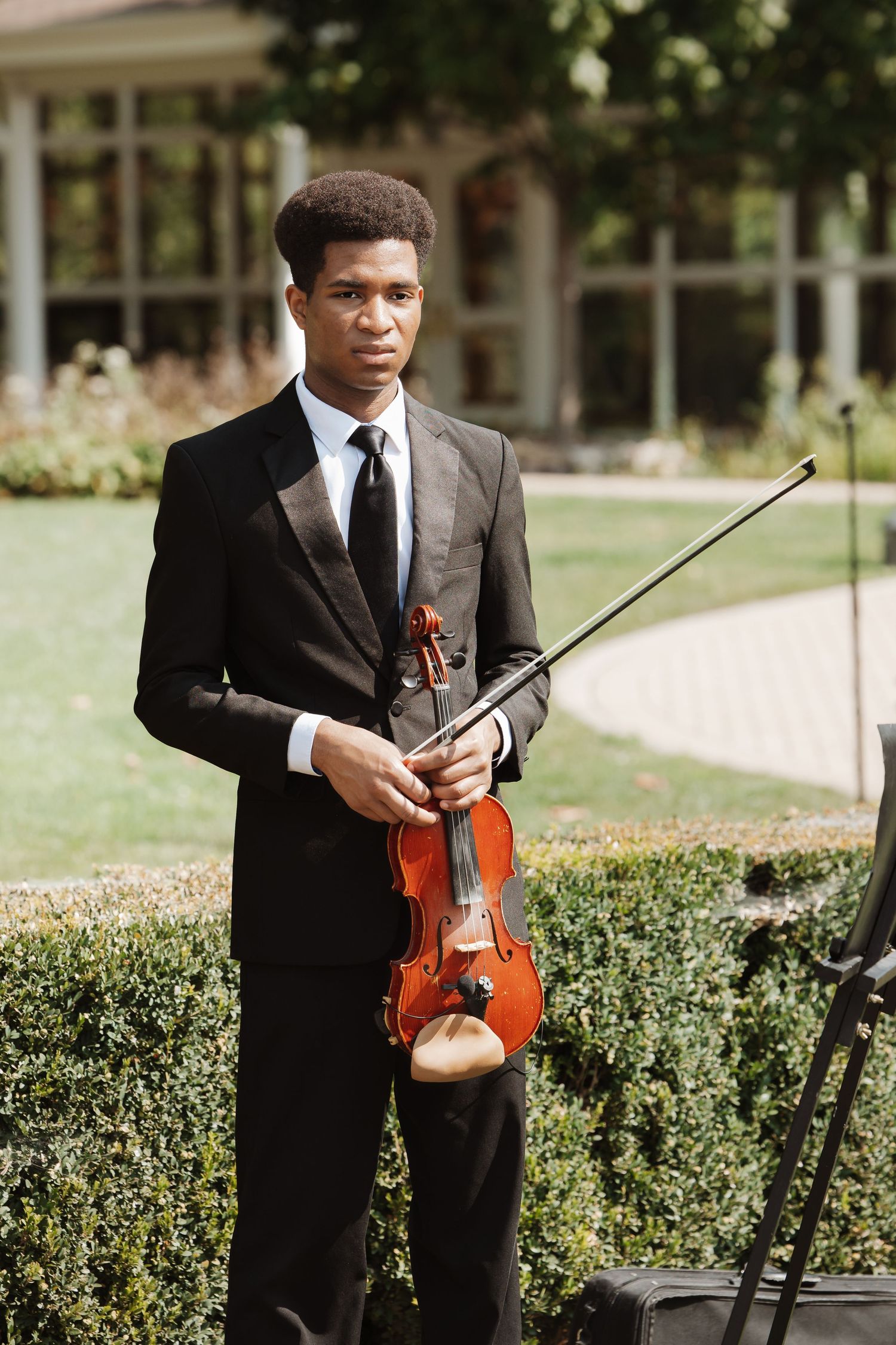 Elegant violinist in black suit holds violin and bow at outdoor wedding venue garden.