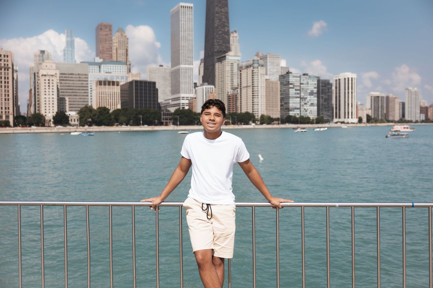 Young man in white shirt and shorts leaning on railing with Chicago skyline and lake behind him.