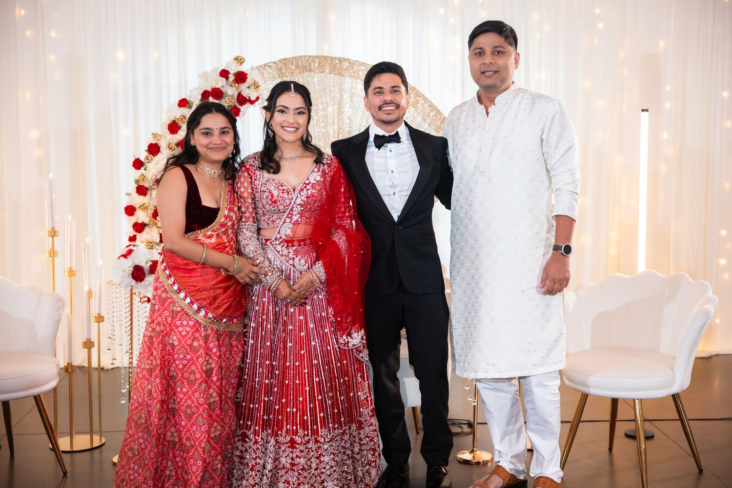 Bride in red lehenga and groom in tuxedo smile with two guests at festive wedding celebration.