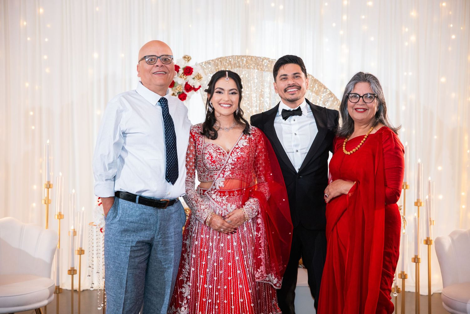 Bride in red lehenga and groom in tuxedo pose with two guests at a fairy-lit wedding venue.