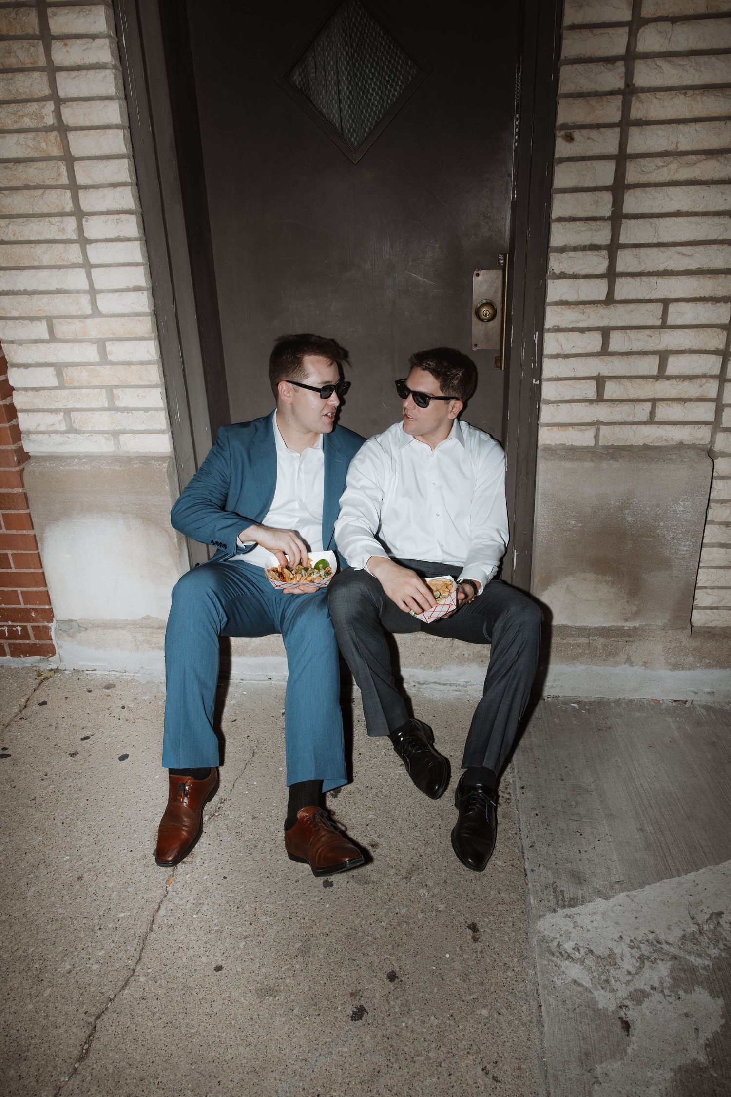 Two men in suits and sunglasses sitting on steps outside EZ Inn, sharing food and conversation.