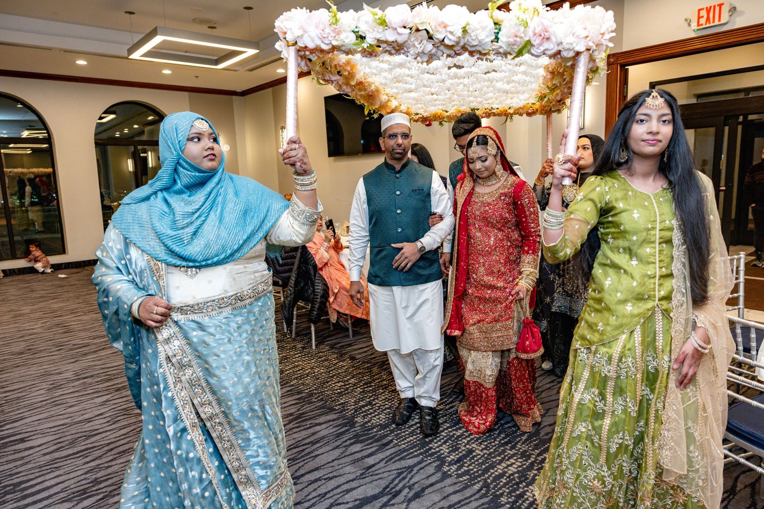 South Asian bride in red lehenga escorted under floral phoolon ki chaadar during wedding procession.