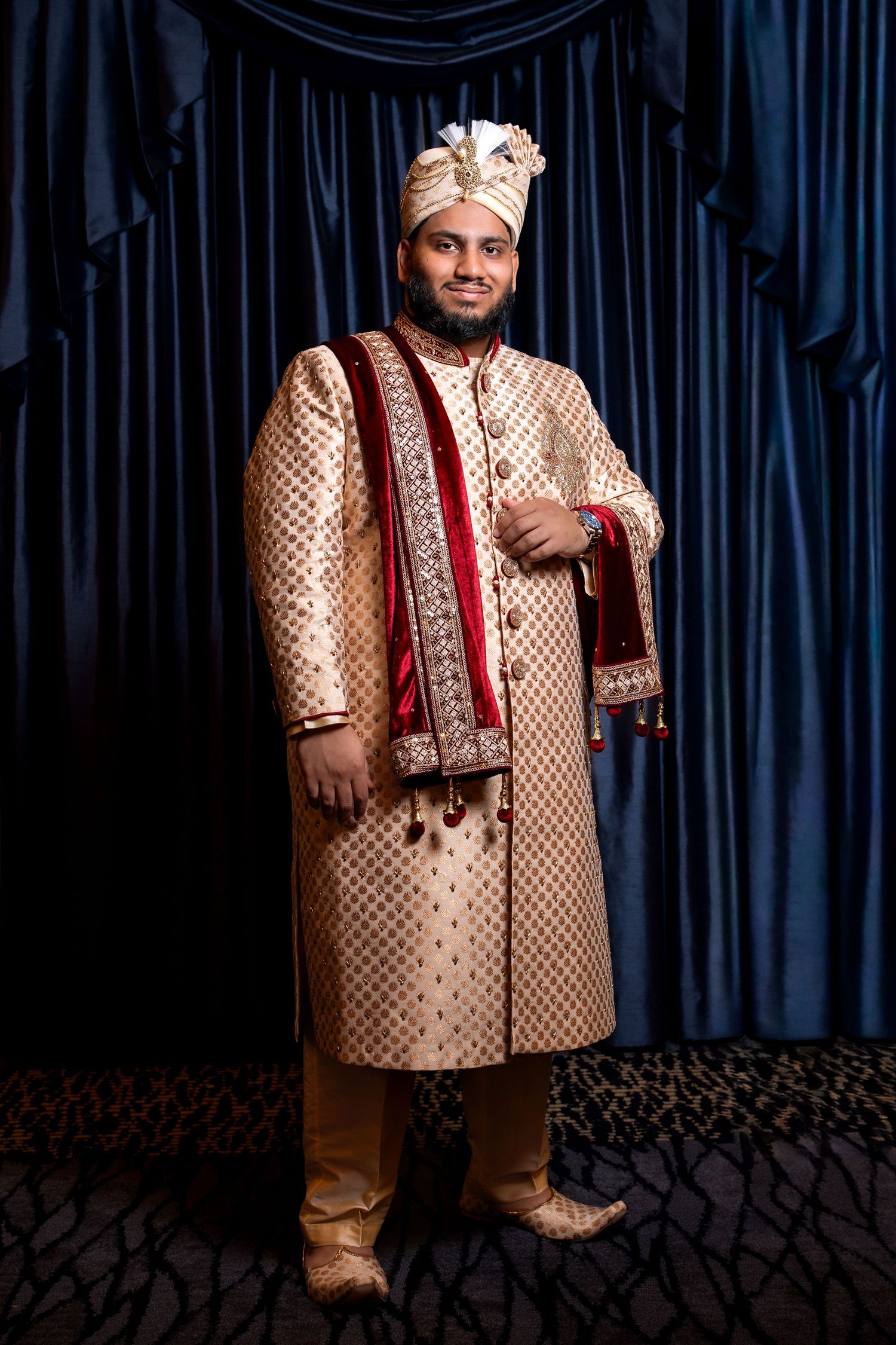 Groom in traditional gold sherwani and turban standing in full-length pose before blue velvet curtains.