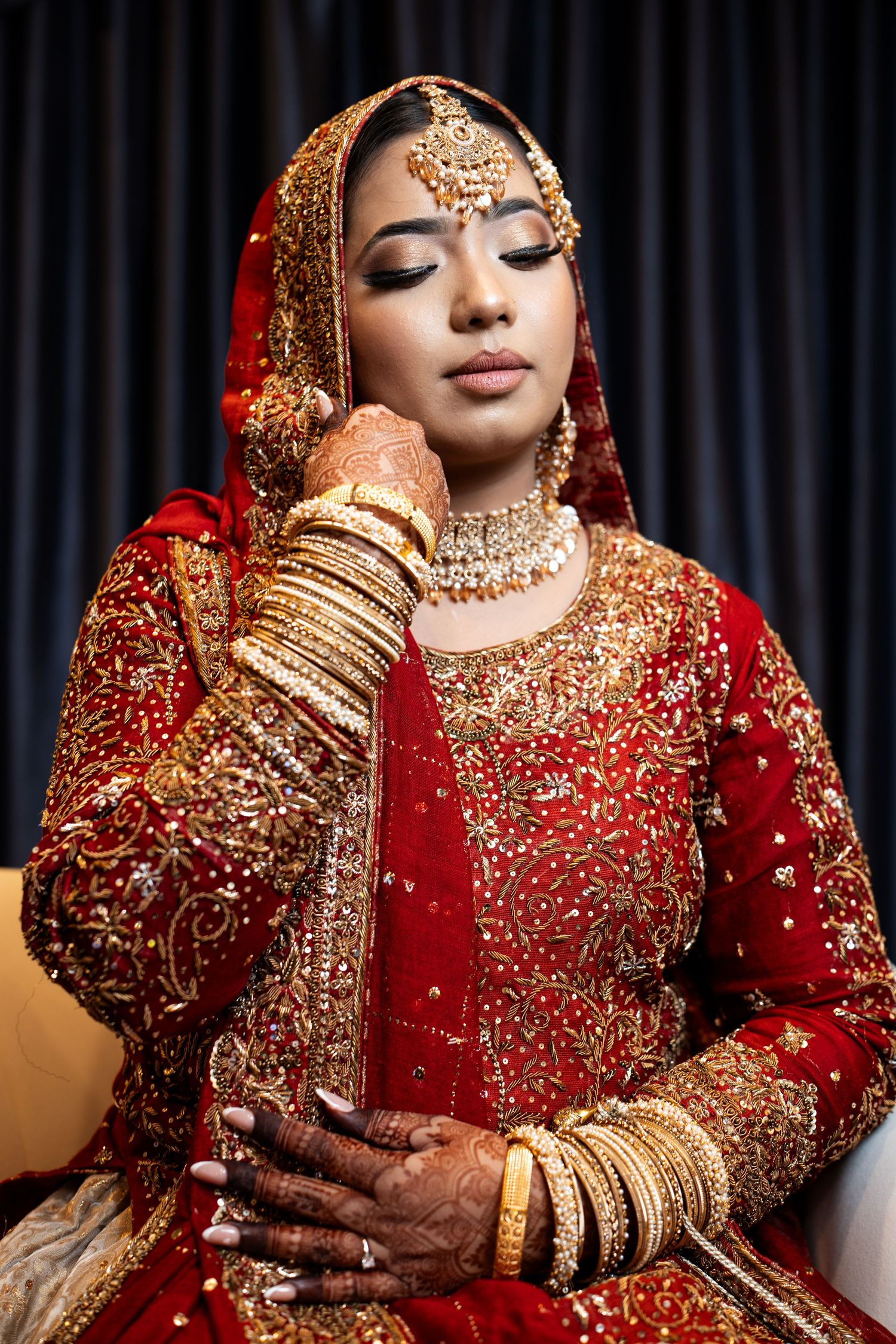 South Asian bride in red lehenga with gold embroidery and bangles poses with eyes closed.