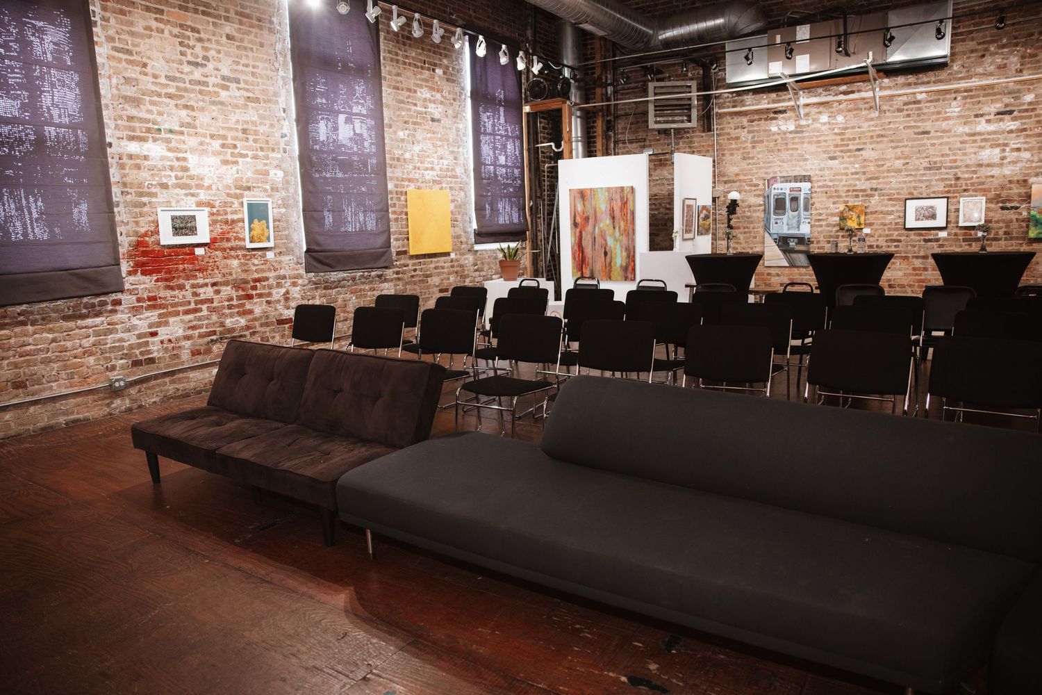 Industrial loft event venue with exposed brick walls, black folding chairs, modern sofas, and art displayed on walls.