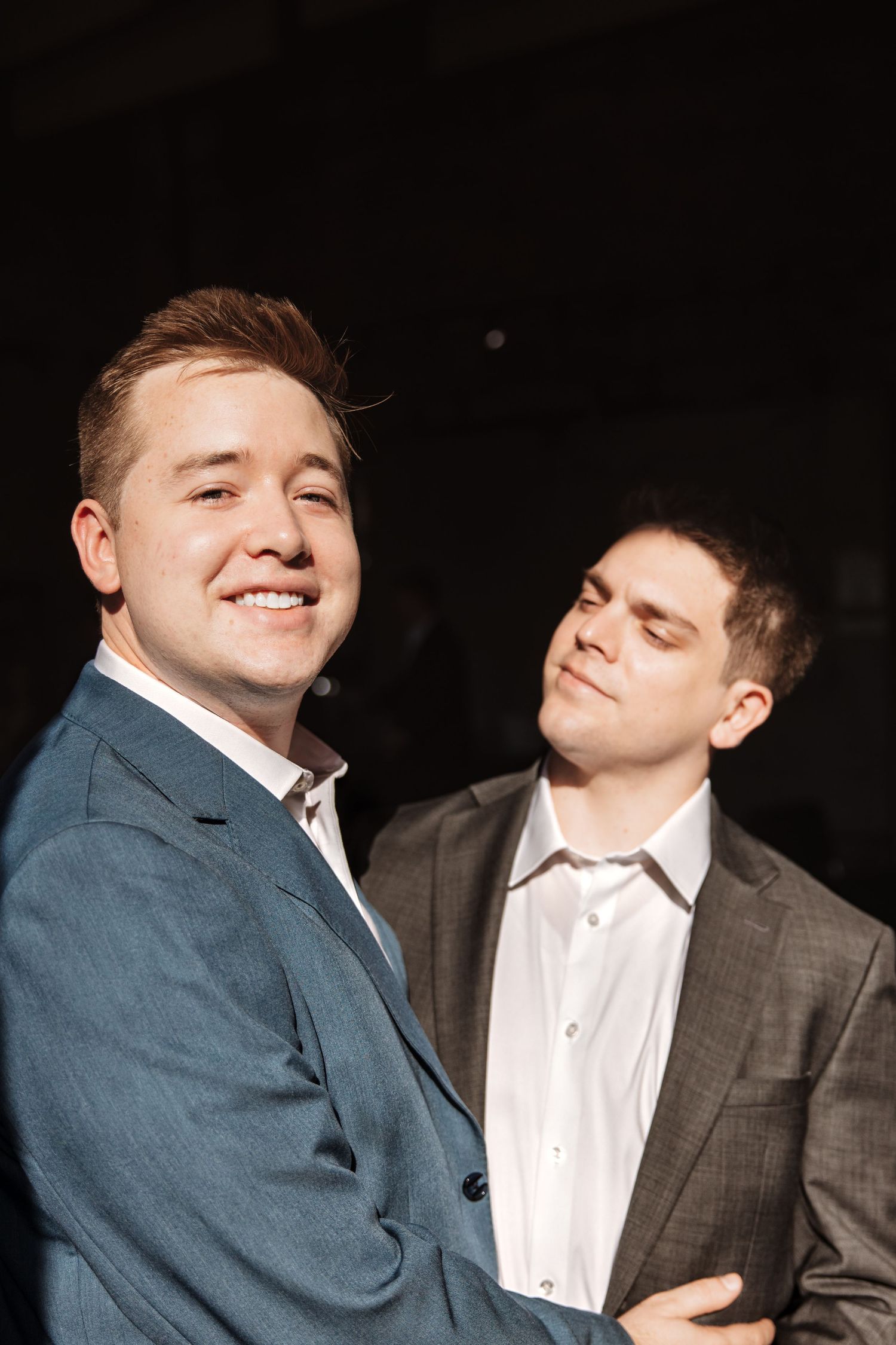 Two young men in blue and gray suits posing playfully together against a dark background.