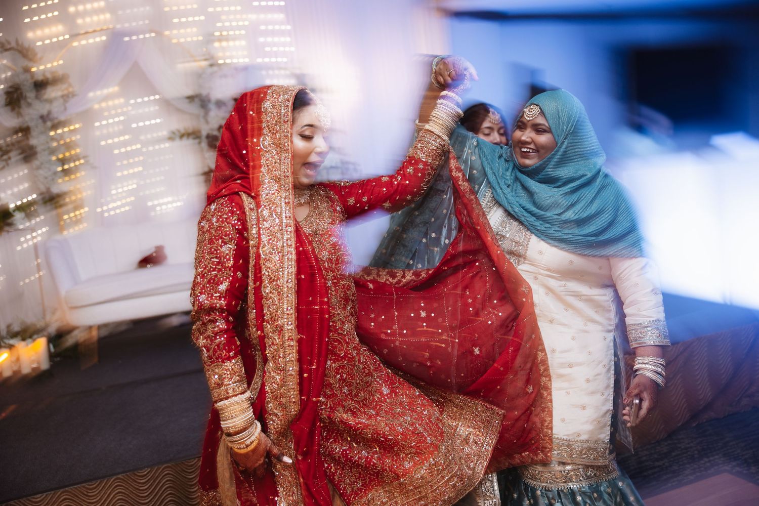Bride in red bridal lehenga dancing joyfully with woman in blue hijab at Indian wedding reception.