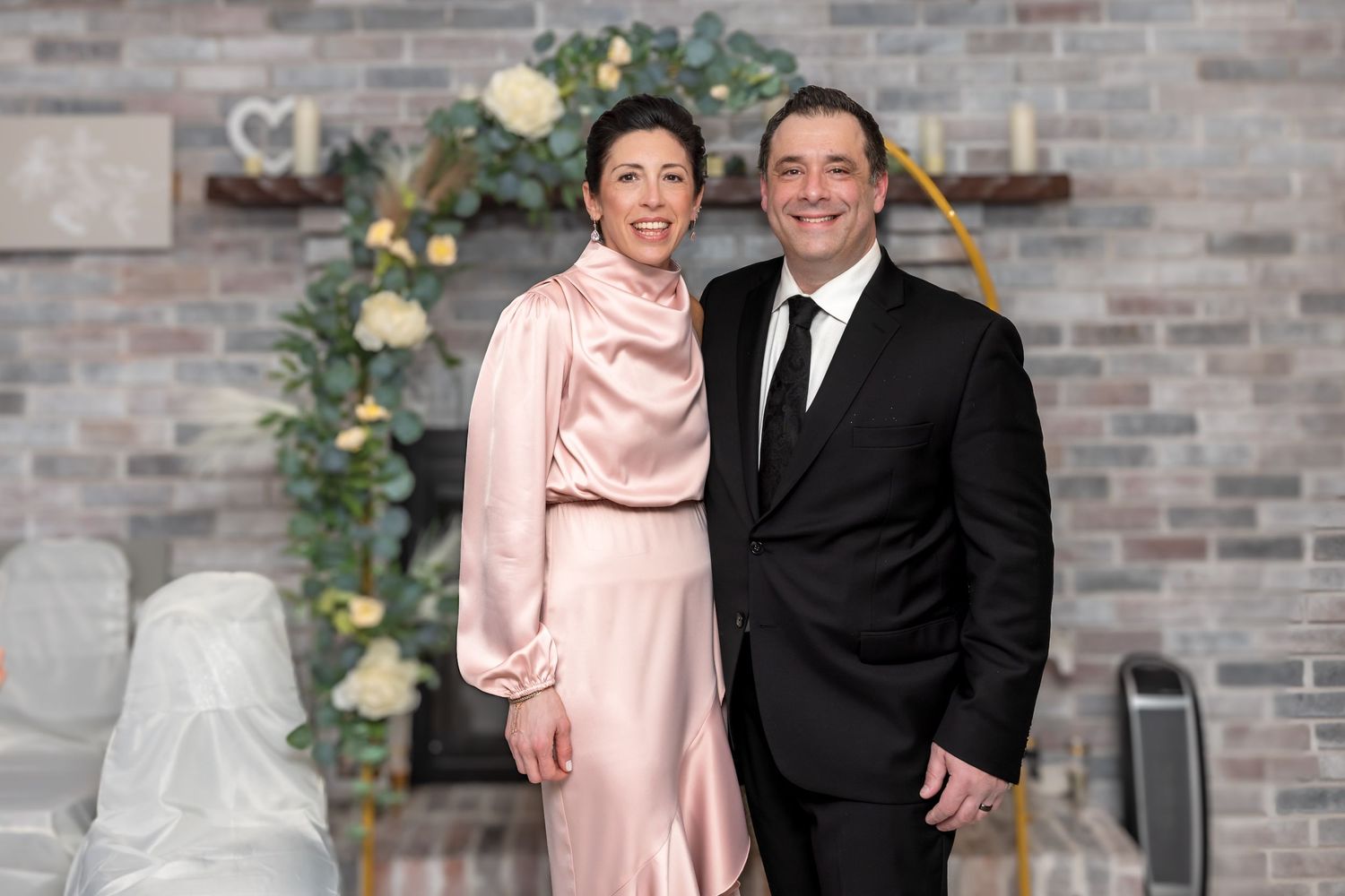 Elegant couple posing together at wedding venue with greenery arch and brick wall background.