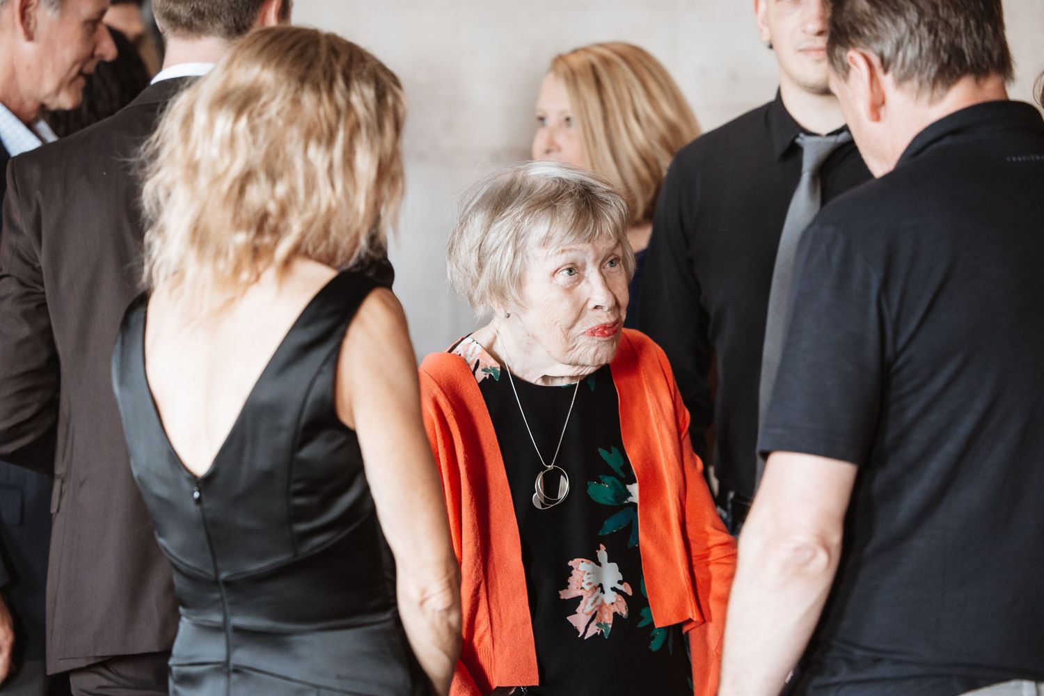Elderly woman in orange cardigan and floral dress socializing at a formal event.