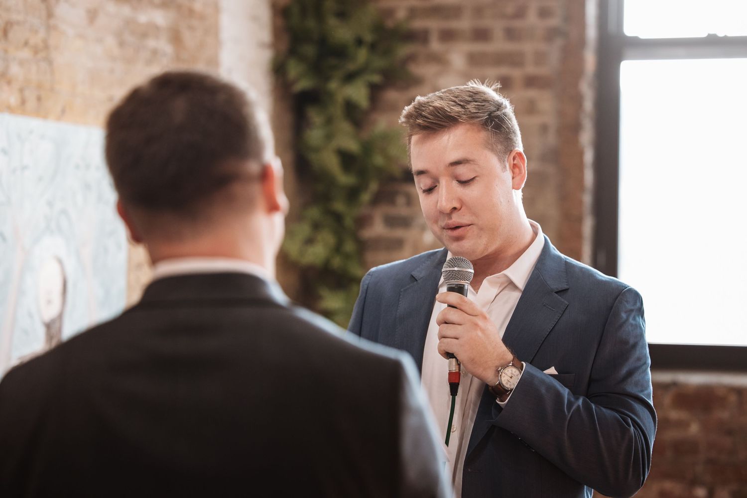 Man in blue suit reads vows into microphone at an intimate indoor ceremony.