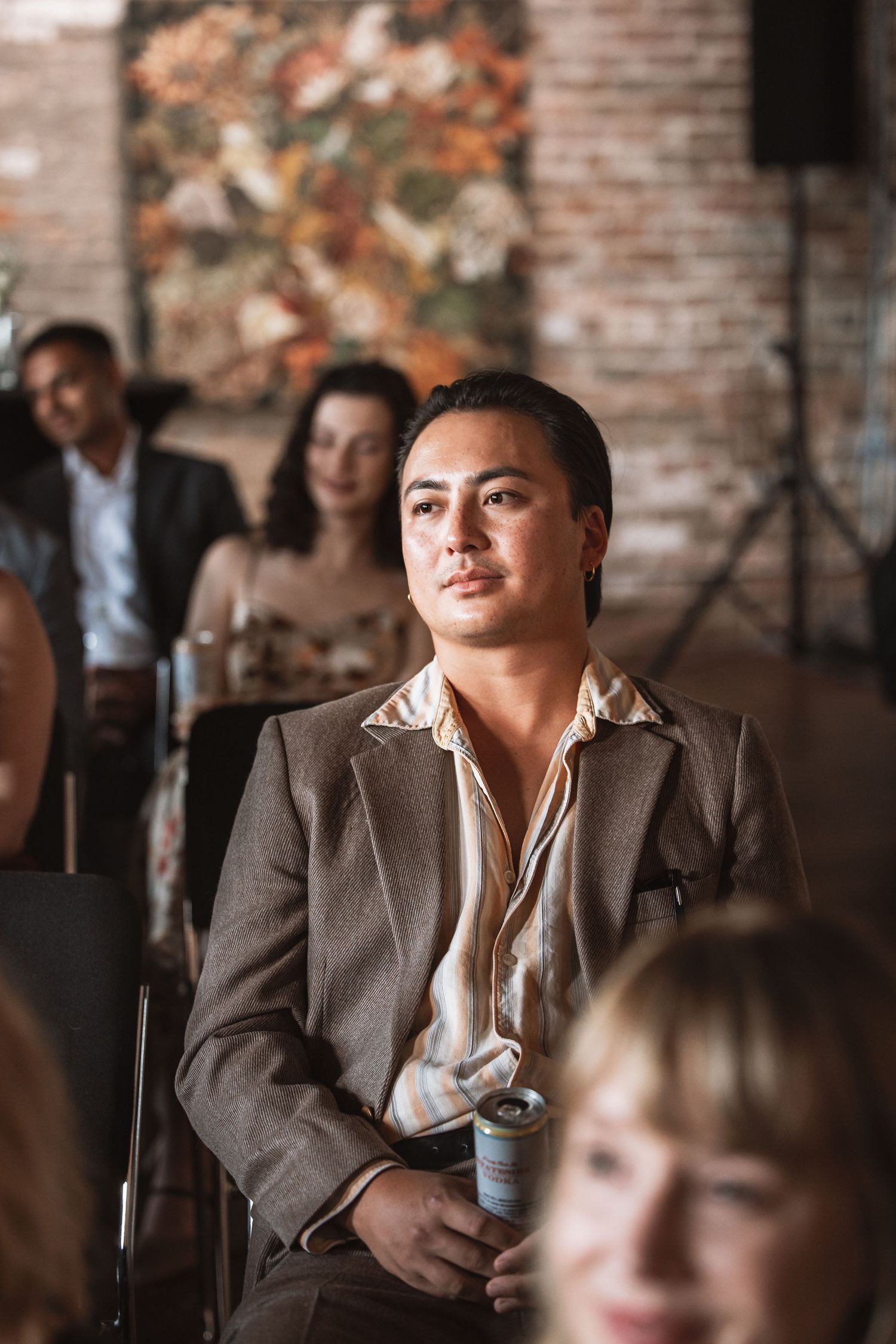 Young man in brown suit holds a drink while attentively watching a presentation.