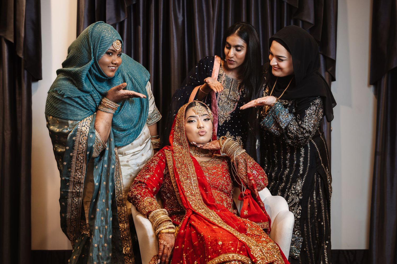 Three women adorning a bride in traditional red Indian bridal outfit with gold embroidery and jewelry.