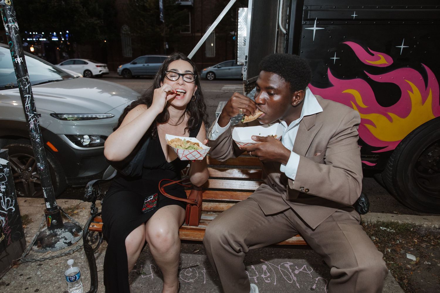 Two guests in formal attire enjoying street food together on a bench at an outdoor evening event.